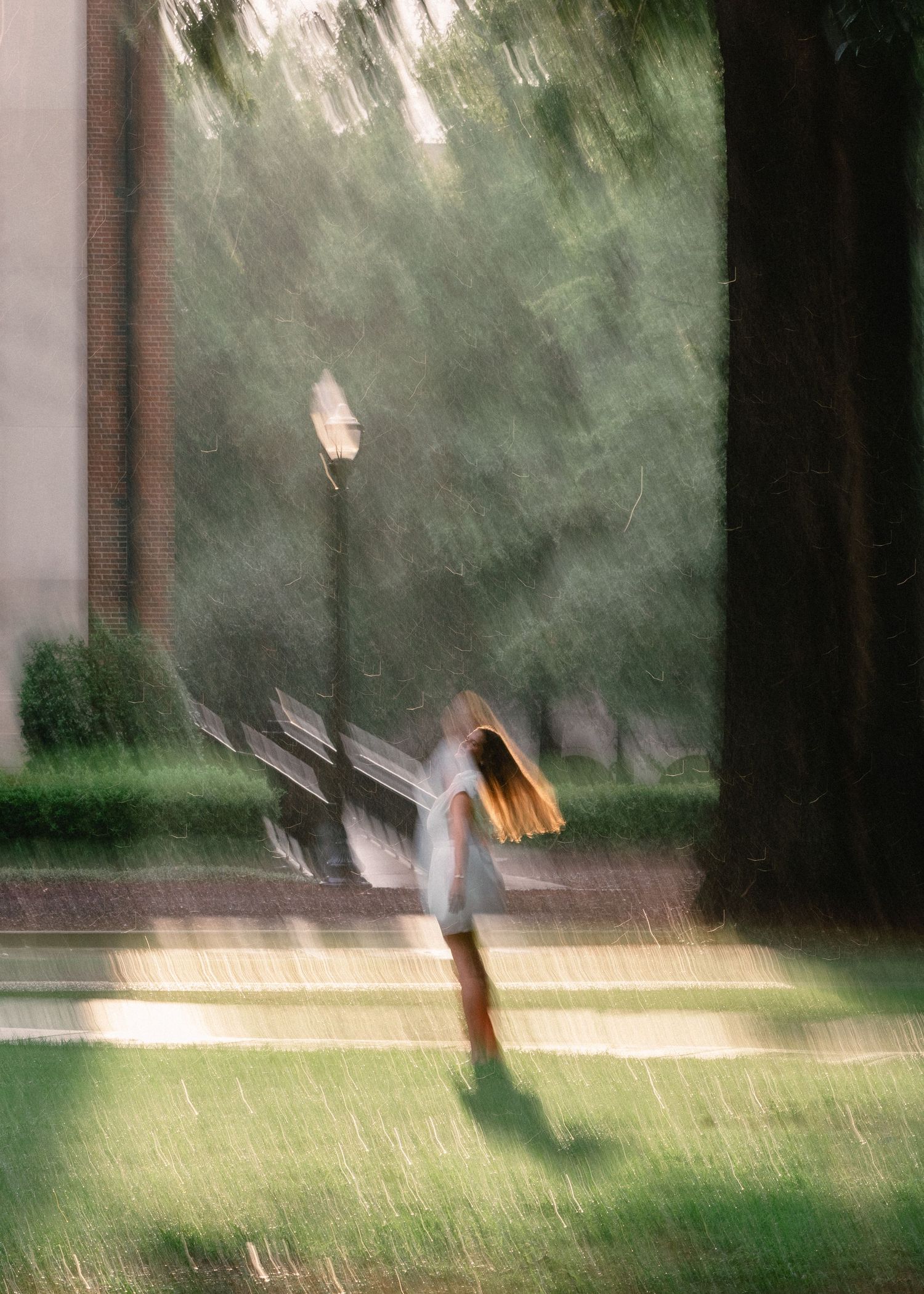 Blurred motion of graduate dancing in heavy rain on sunlit campus pathway.