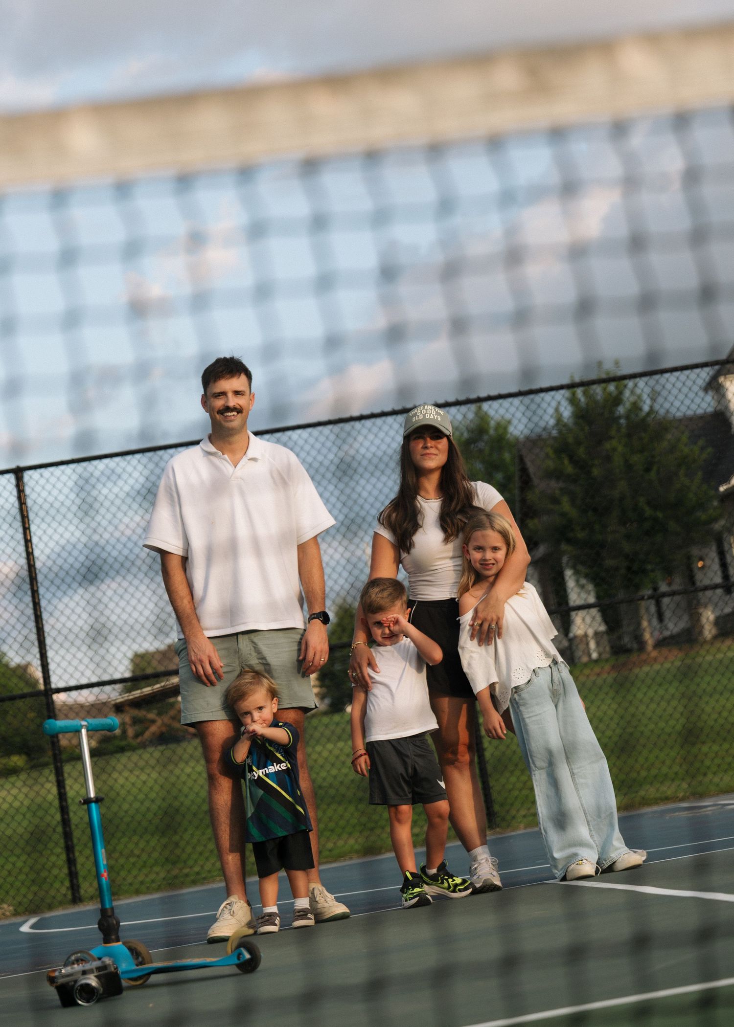 A family of five stands together on a tennis court, viewed through the net on a cloudy evening.