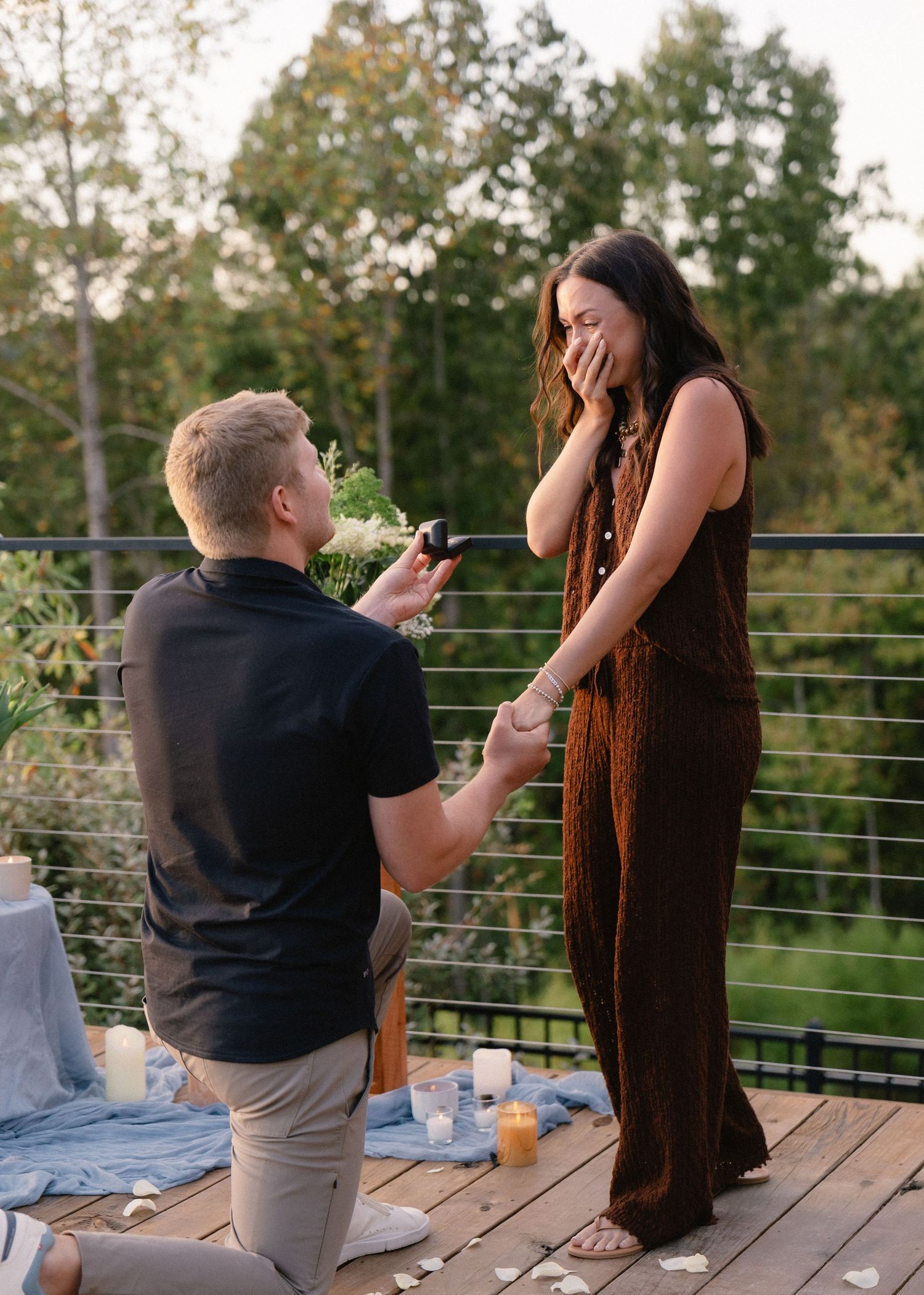 Man kneeling on a wooden deck holds out a ring as a surprised woman covers her mouth during a sunset marriage proposal.