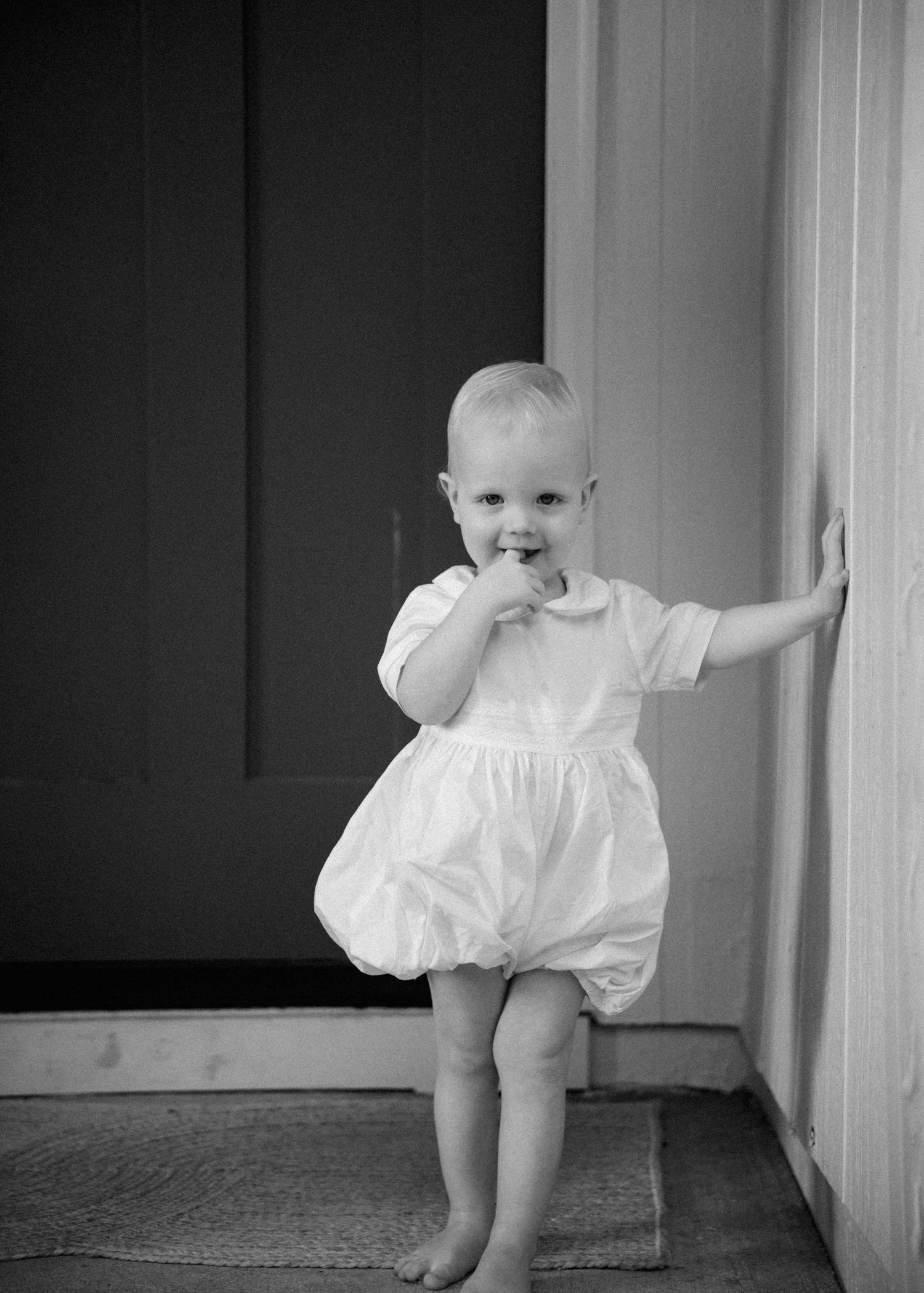 Black and white photo of a barefoot toddler in a white bubble romper standing on a doormat, hand on wall and fingers in mouth.
