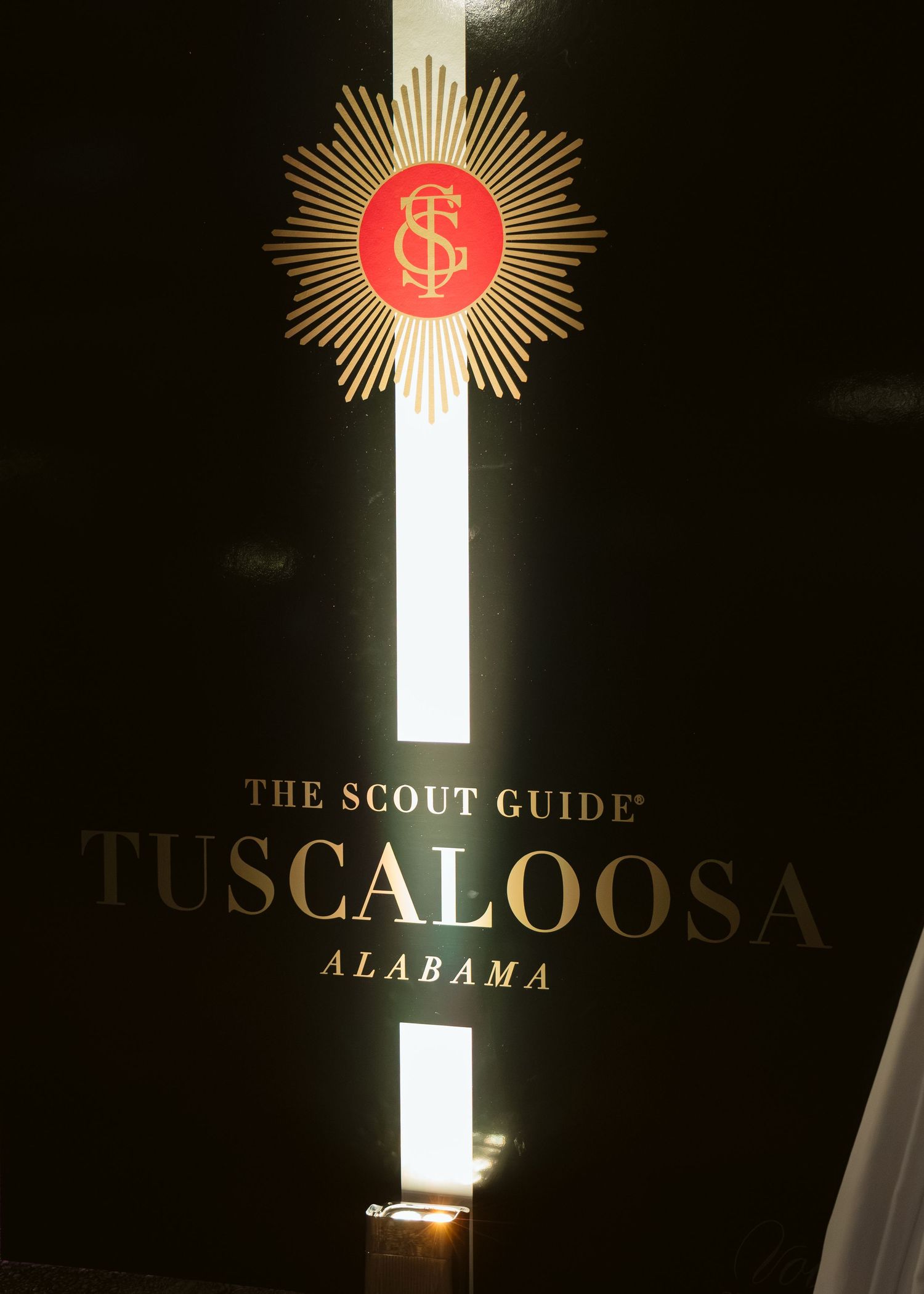 Illuminated black sign with a gold sunburst logo reading The Scout Guide Tuscaloosa, Alabama.