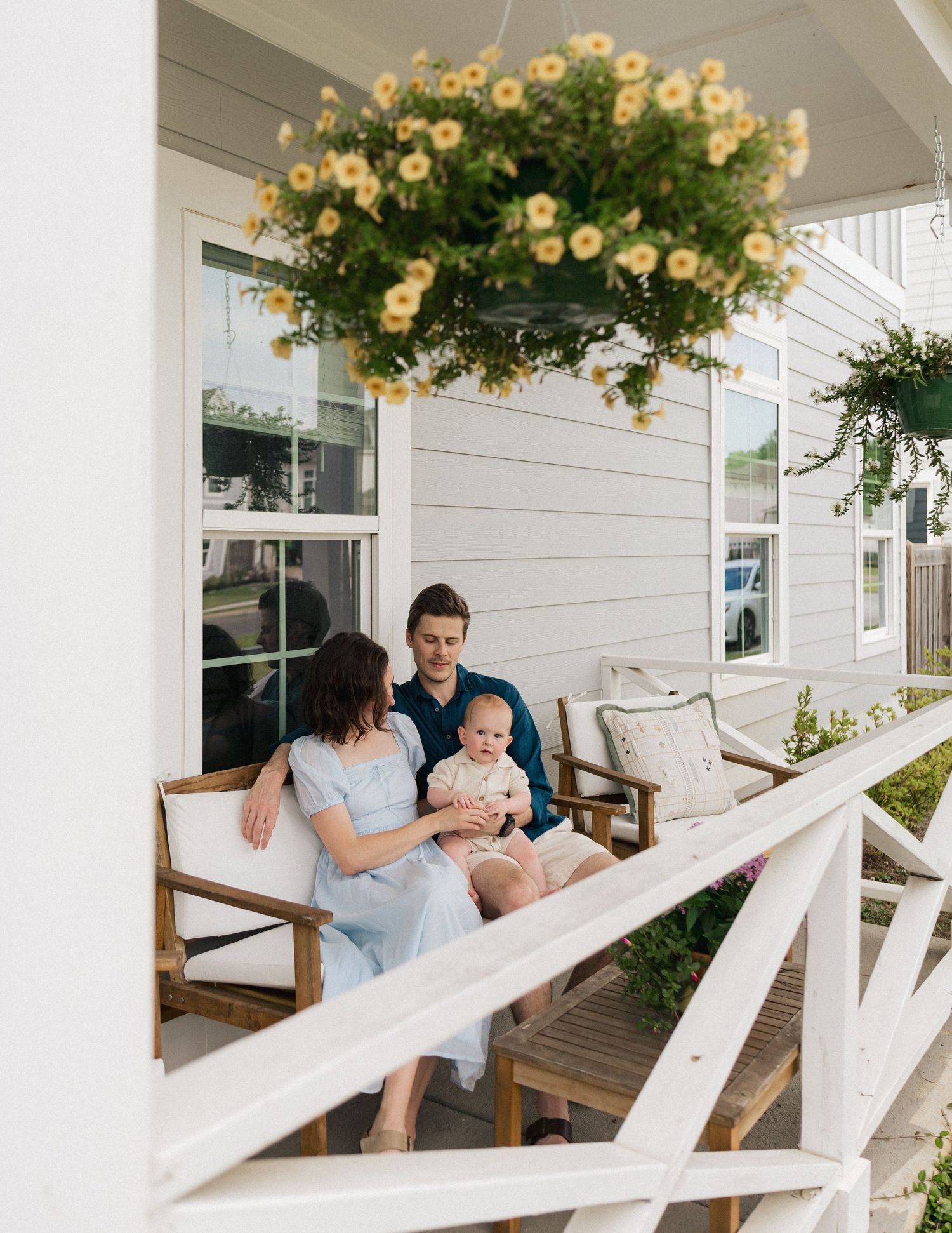 A young family with baby relaxes on a charming front porch with yellow flowers.