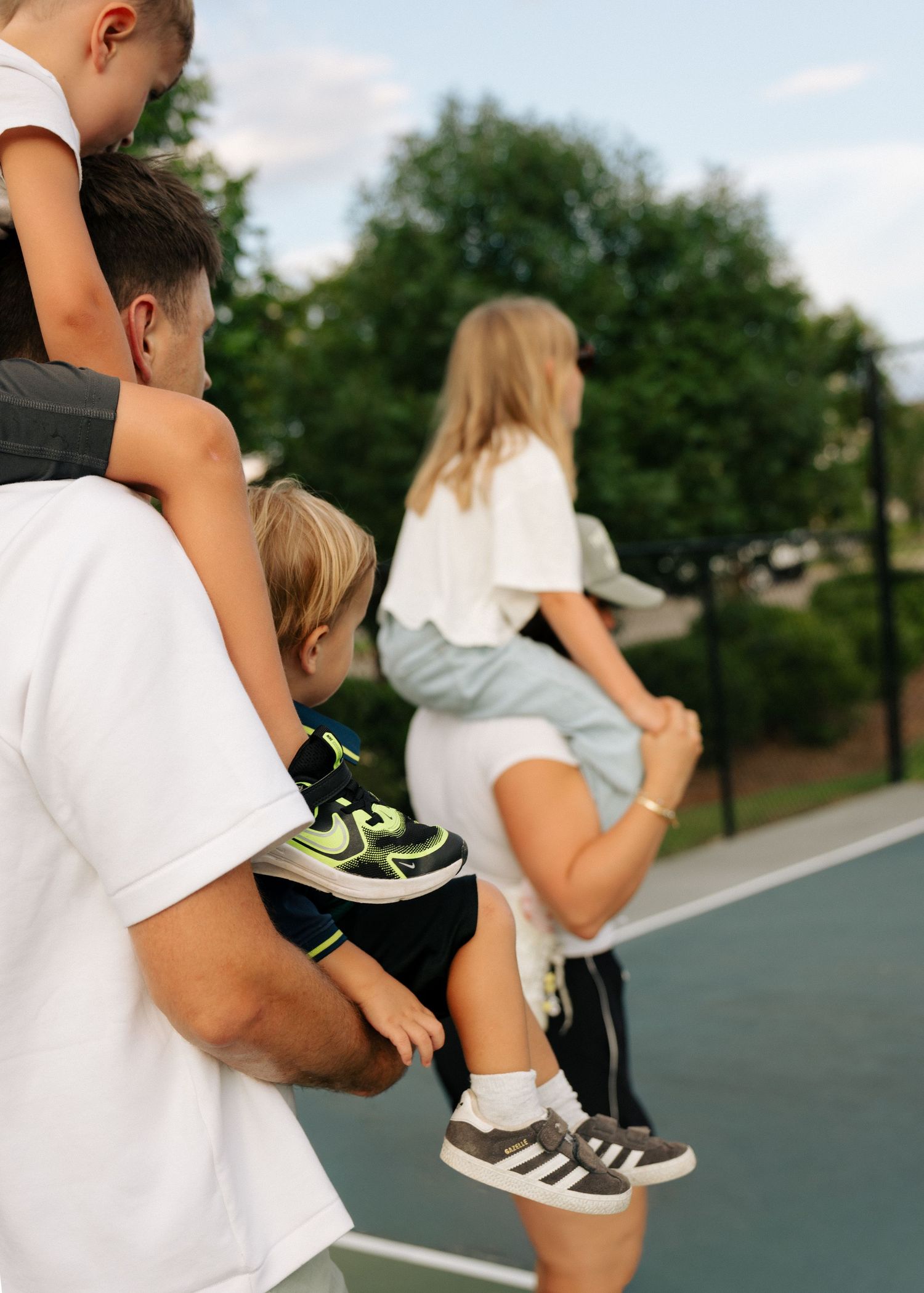 A father carries two young children on a tennis court while a woman stretches nearby on a sunny day.
