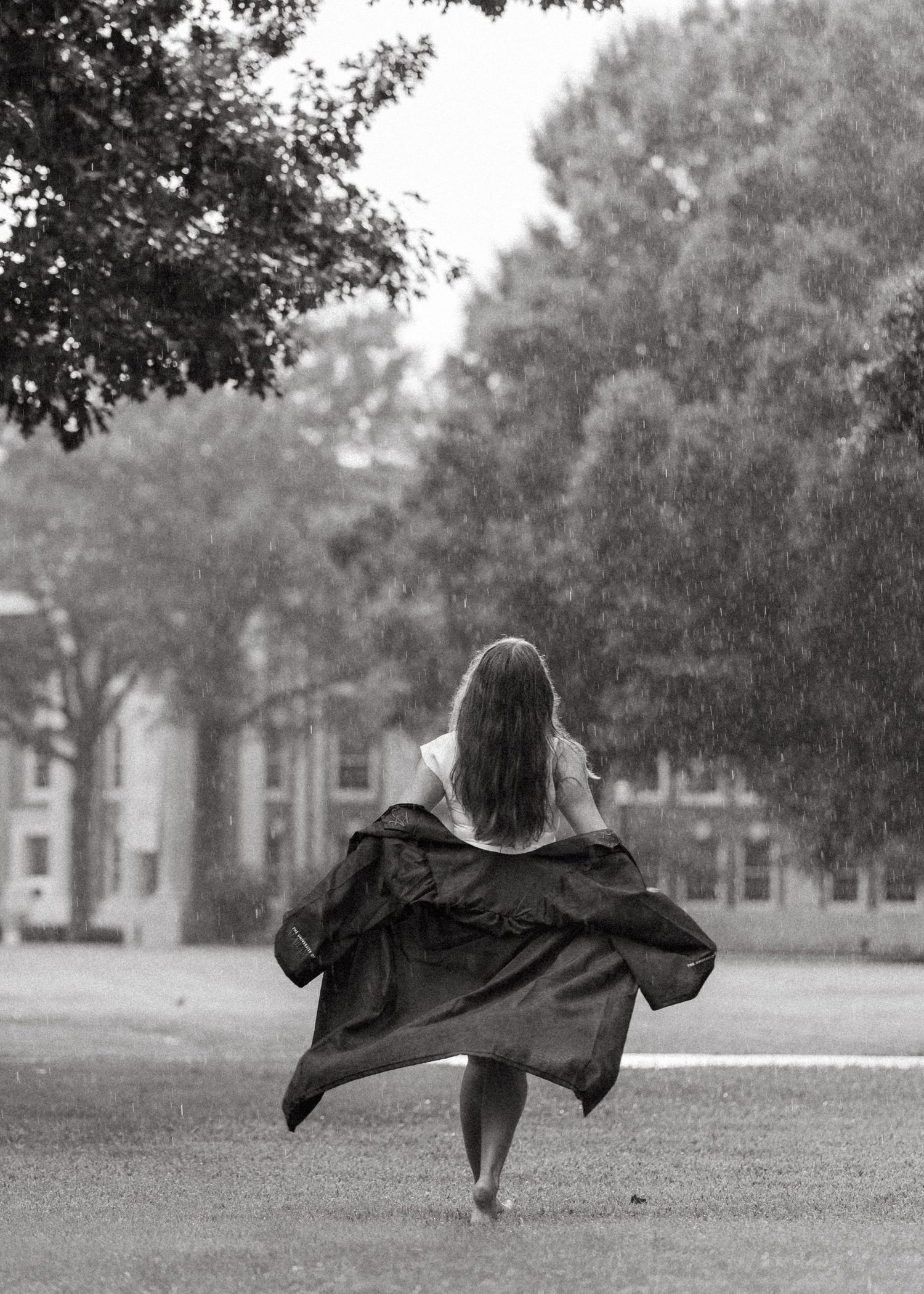 Graduate runs barefoot in rain wearing black gown on lush green campus lawn.