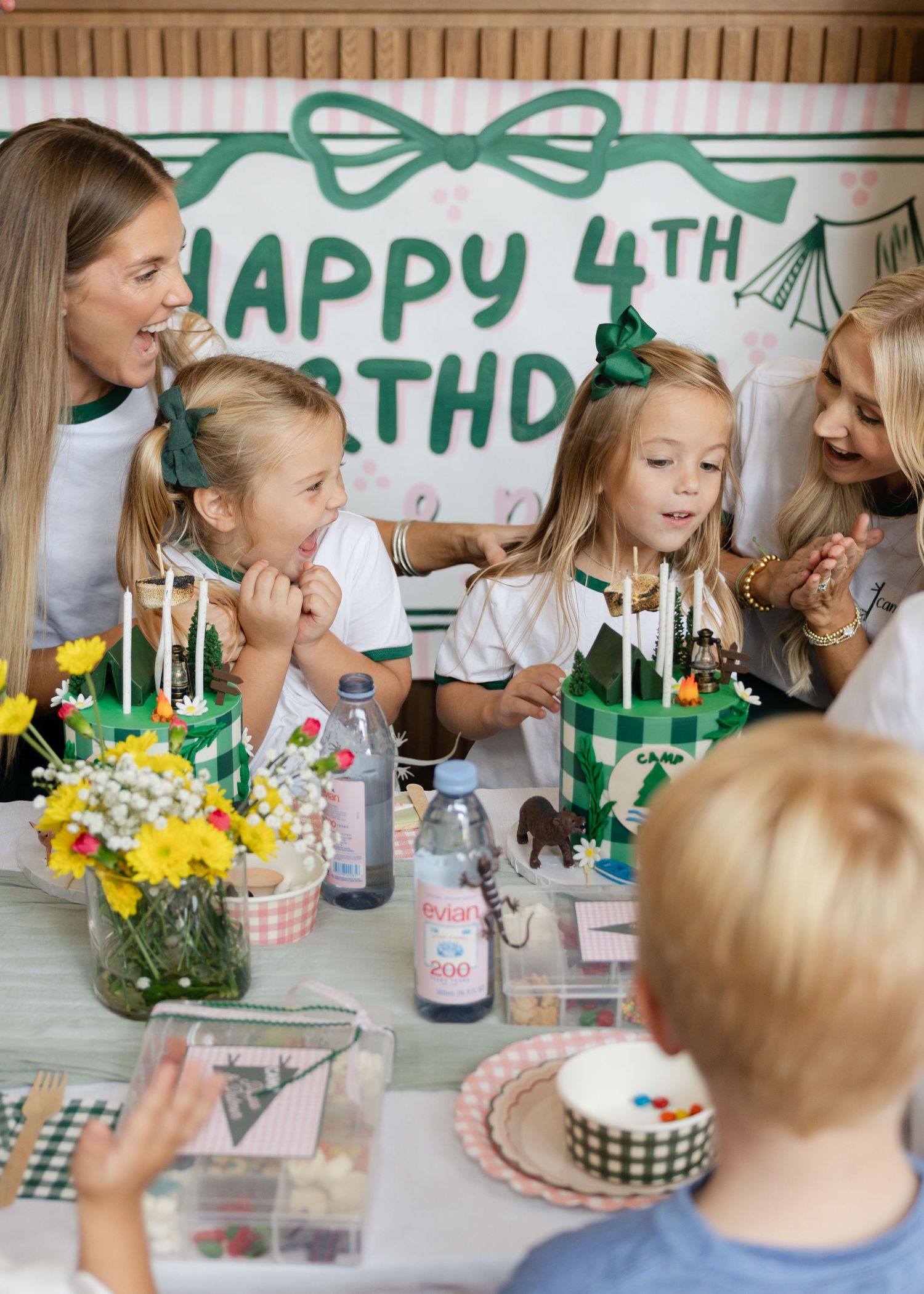 Two mothers and twin girls celebrate their 4th birthday with candles and cake at a camp-themed party.
