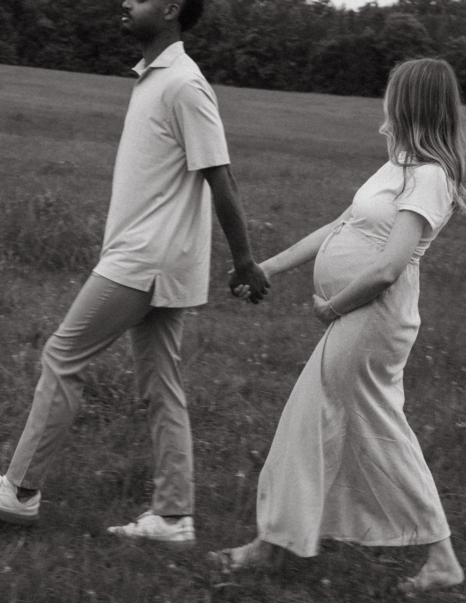 Black and white maternity photo of couple holding hands while walking through an open grassy field.