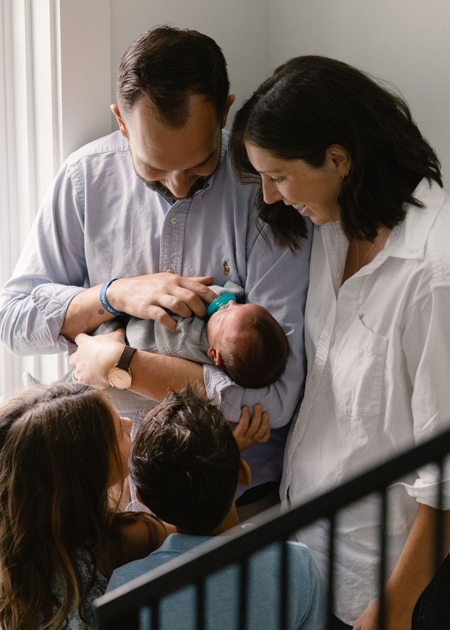Family of four gathered together admiring their newborn baby indoors near staircase.