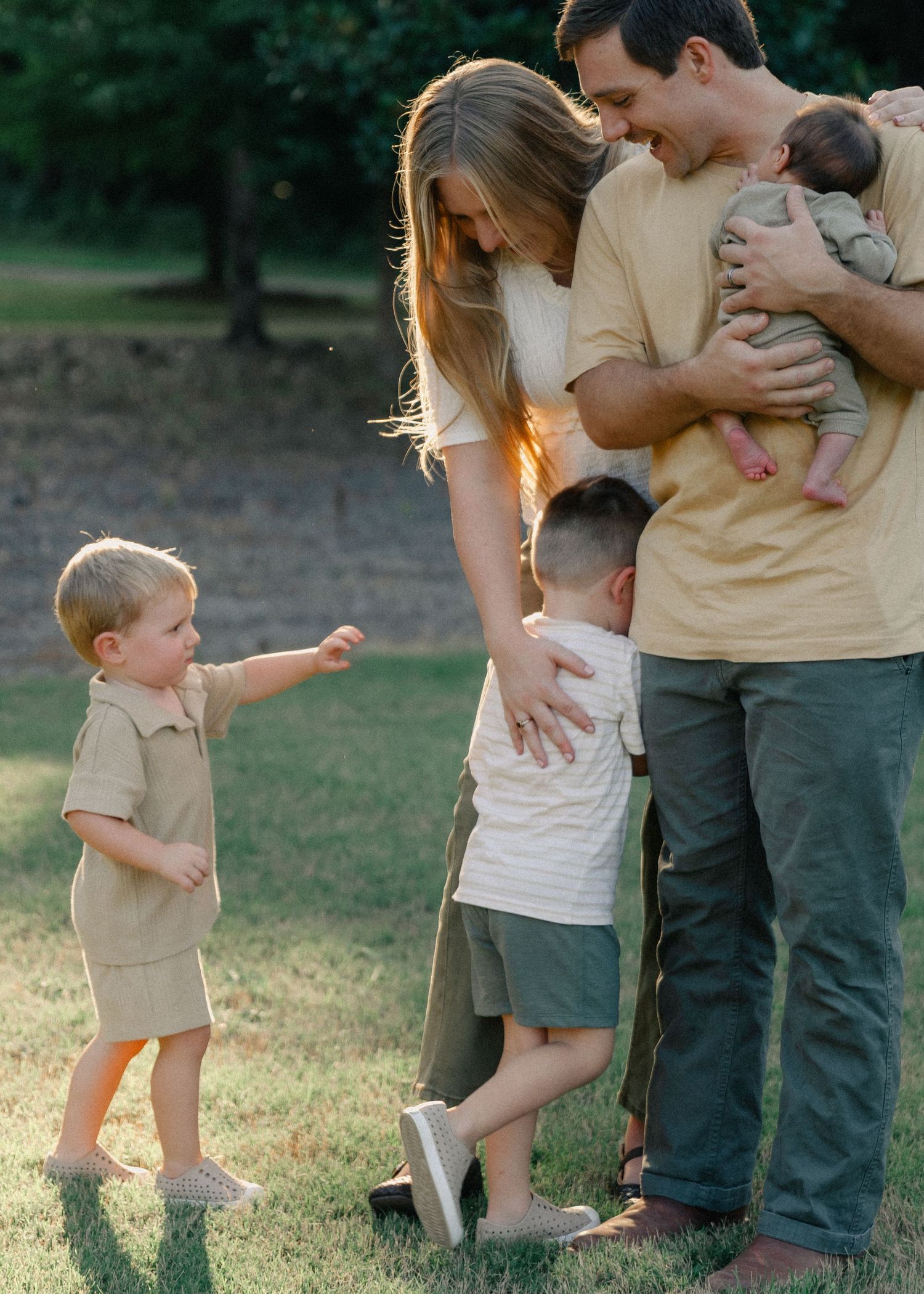 Young family of five outdoors at golden hour, parents holding baby while two toddler boys play nearby.