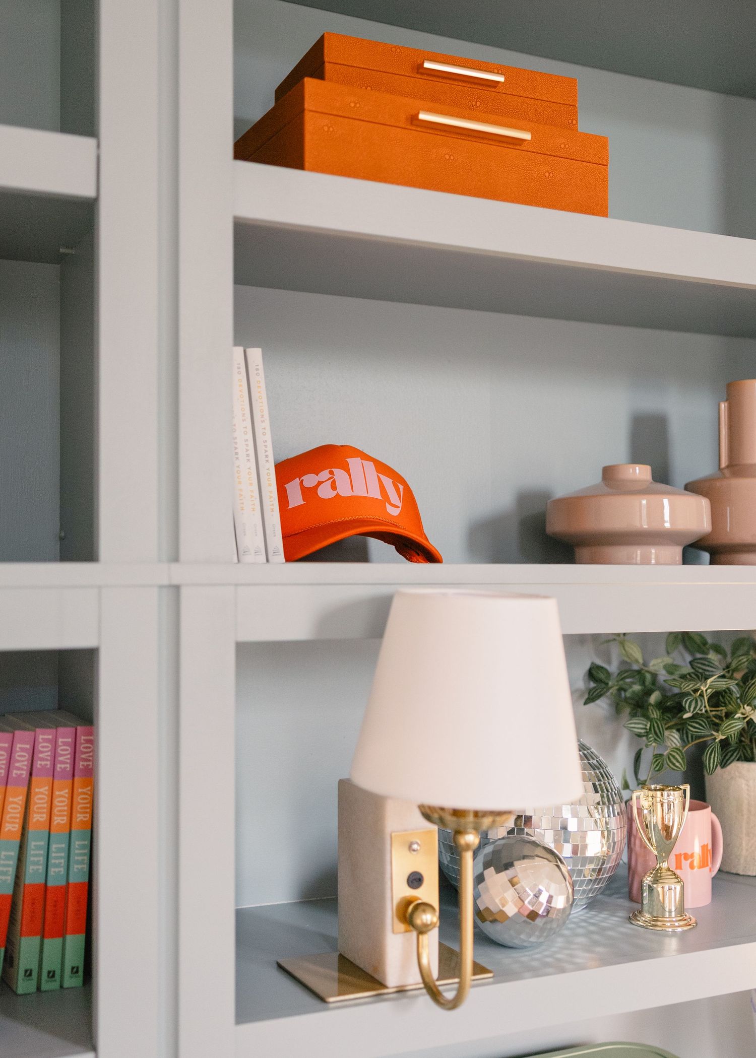 Styled white bookshelf with an orange rally cap, orange storage box, lamp, and decorative accessories.