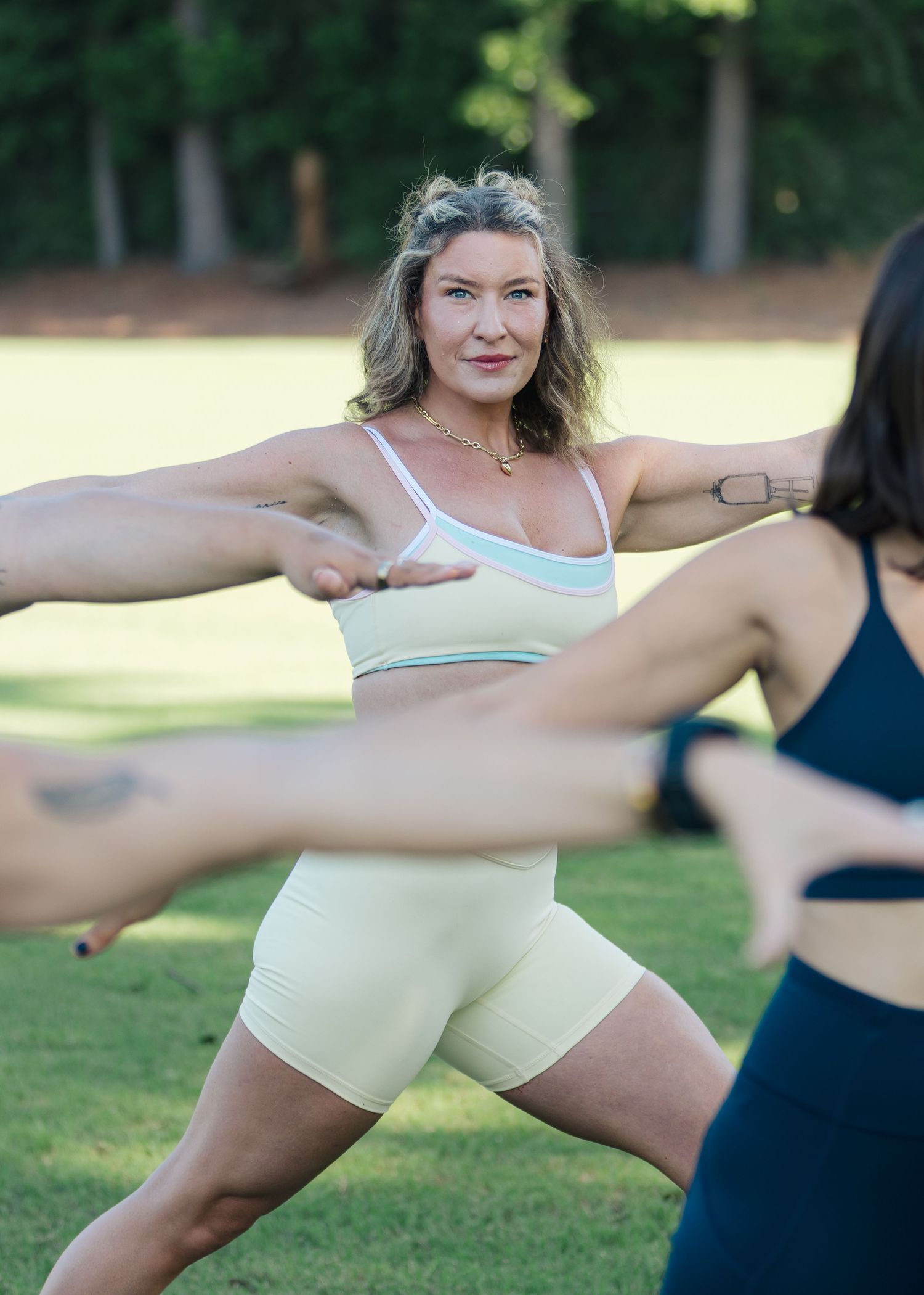 Woman in yellow sports bra and shorts doing a warrior yoga pose outdoors in a green park.
