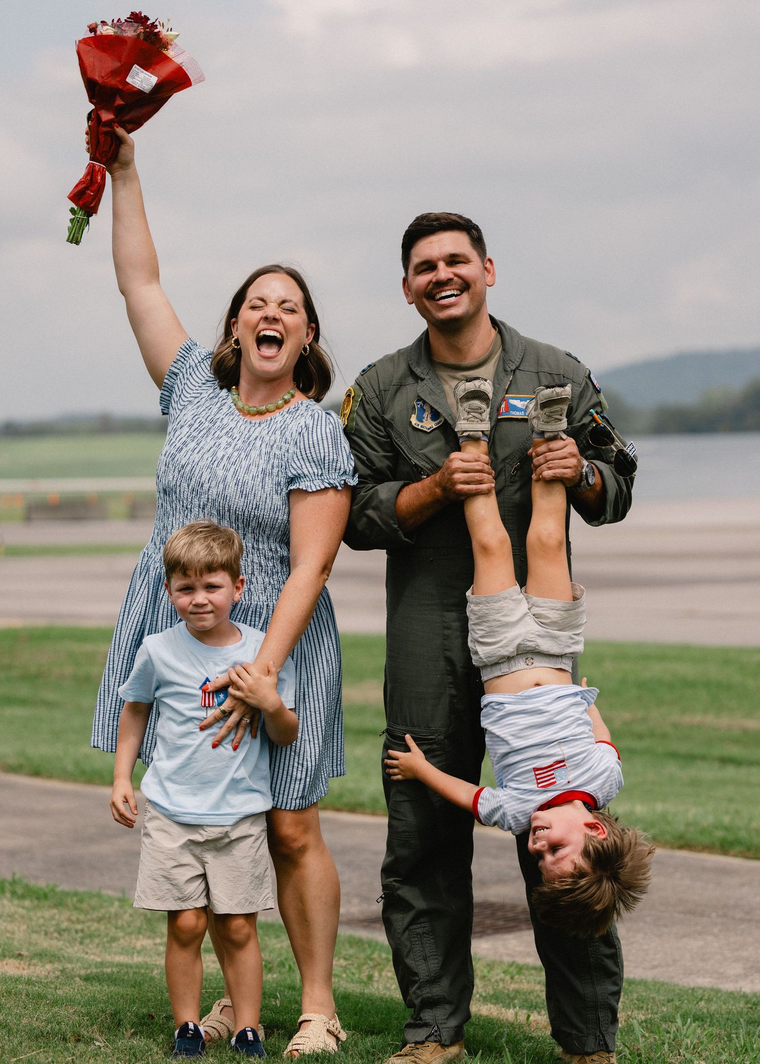 Joyful military family reunites on airfield, mother raising flowers, pilot father holding child upside down with two sons.