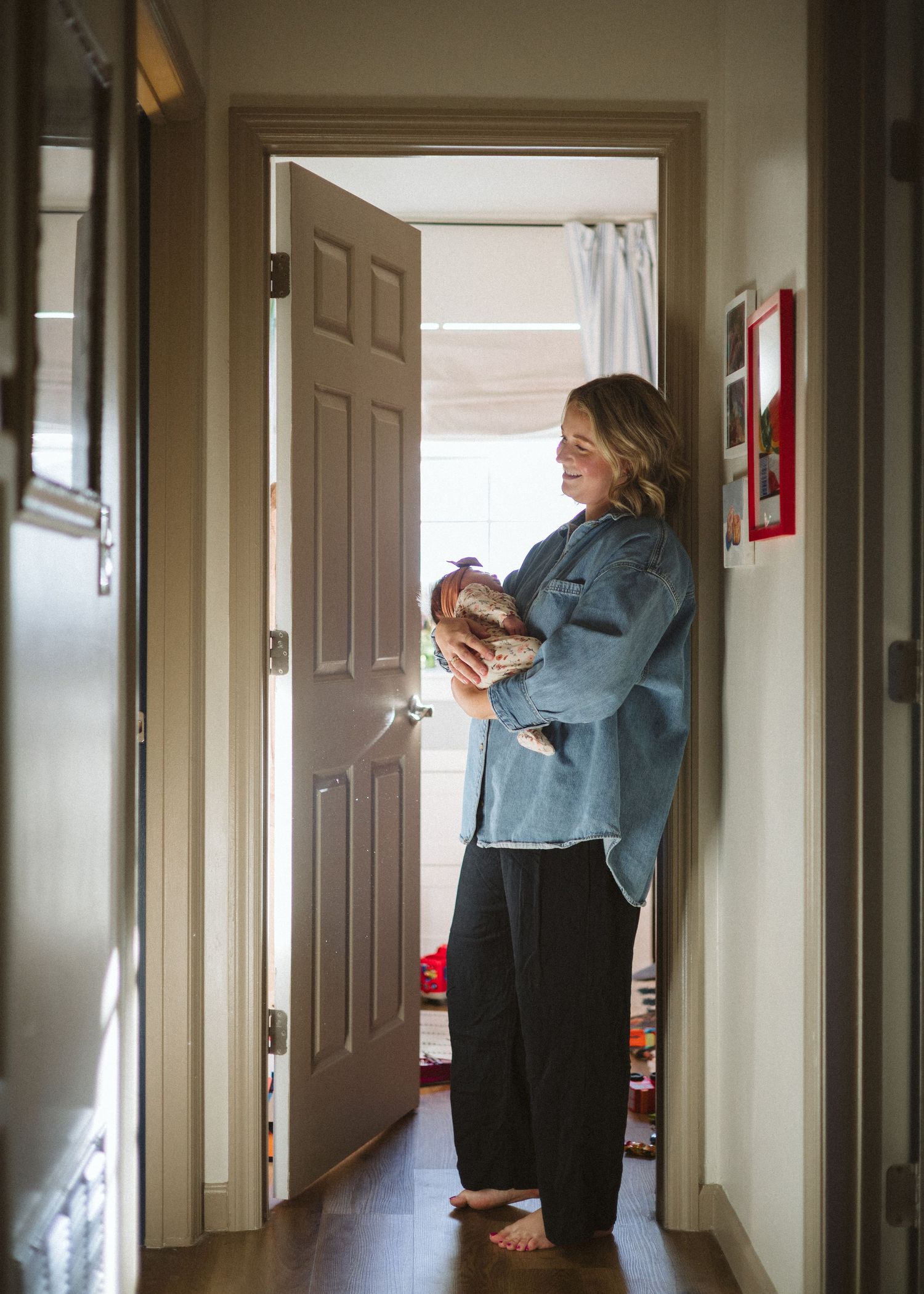 Barefoot woman in a denim jacket and black pants smiles while cradling a sleeping newborn in a sunlit hallway doorway.