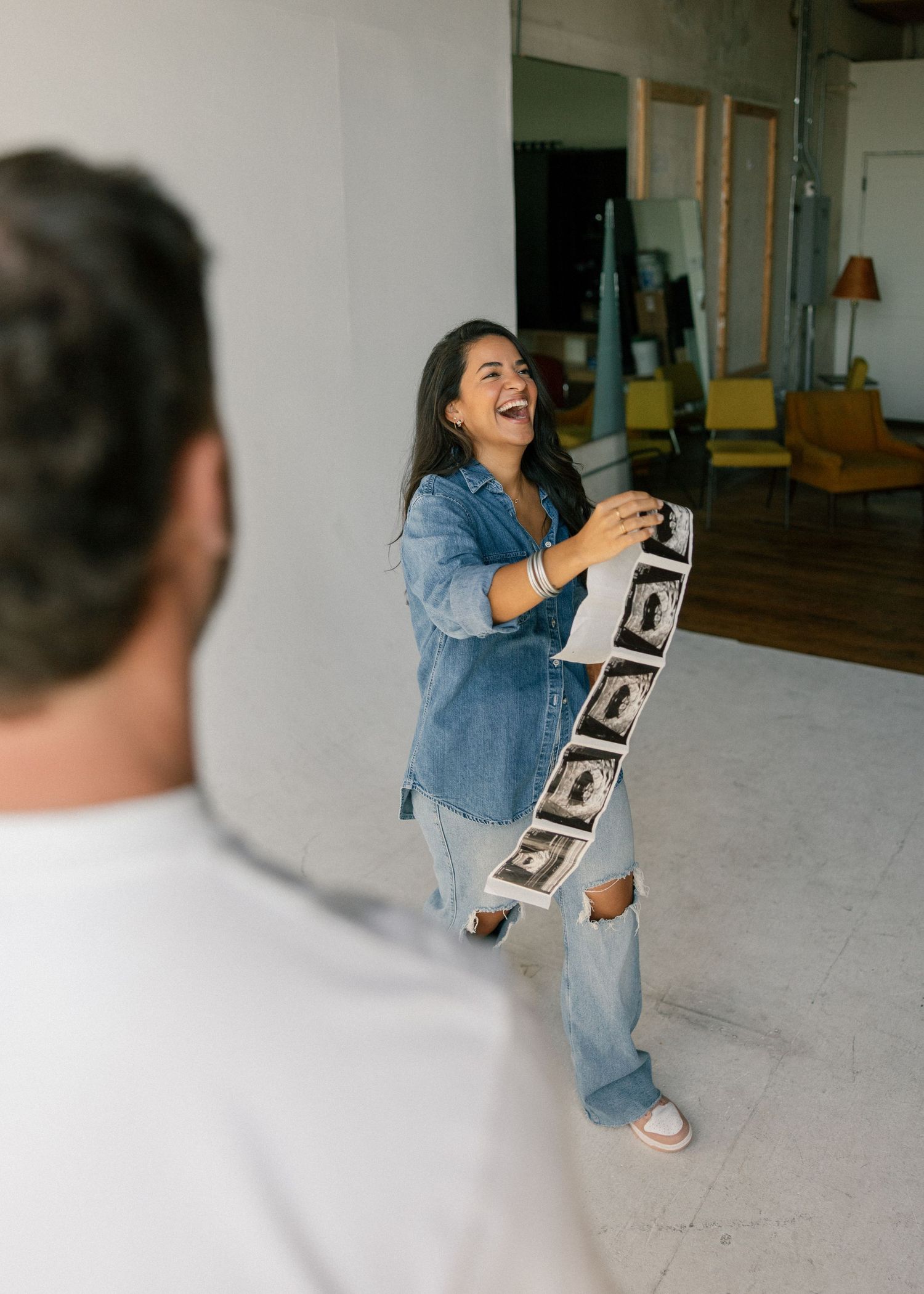Laughing woman in denim jacket holding ultrasound strip photos during a joyful pregnancy announcement shoot.