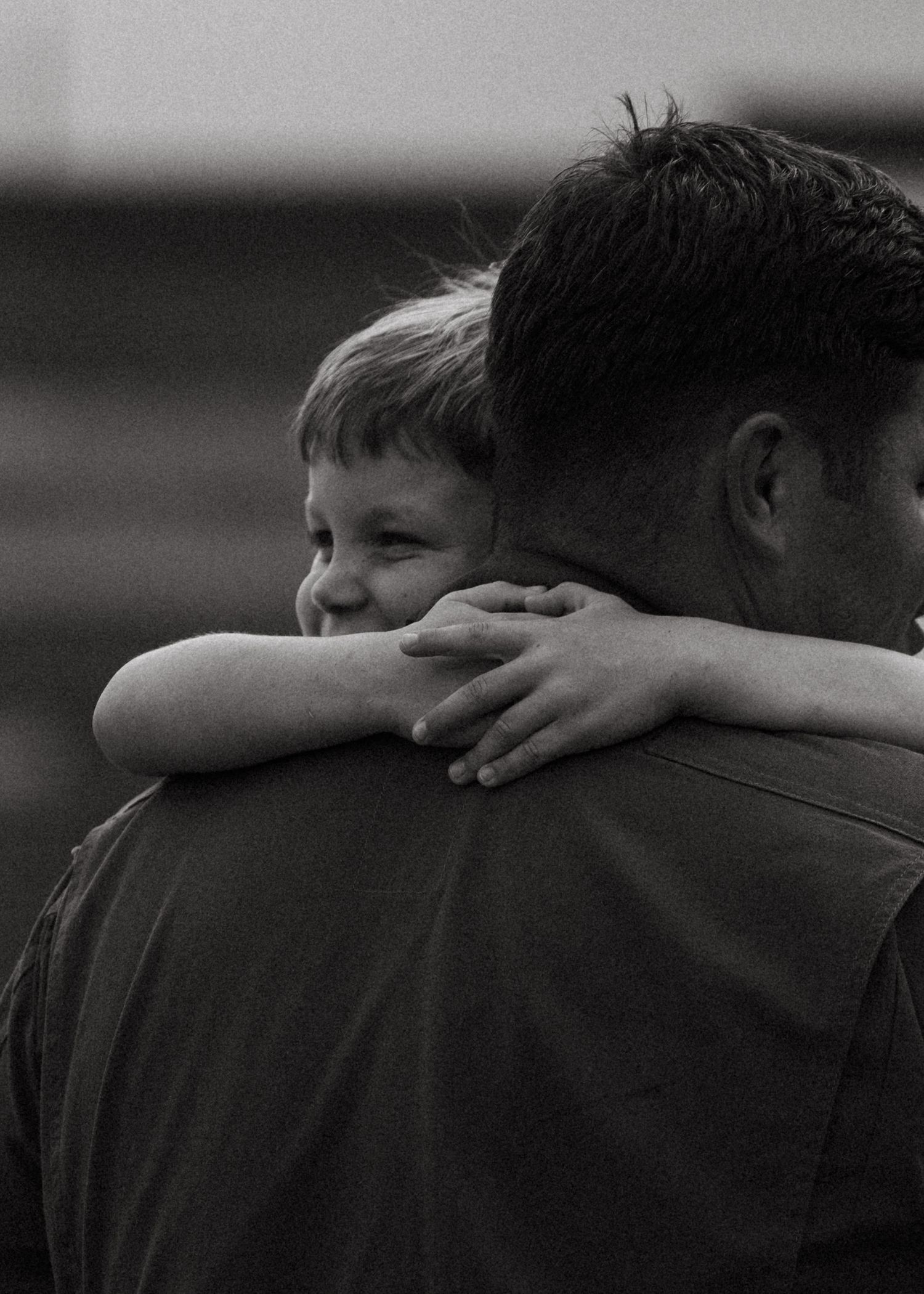 Black and white photo of a young child smiling over a military parent's shoulder during an emotional homecoming hug.