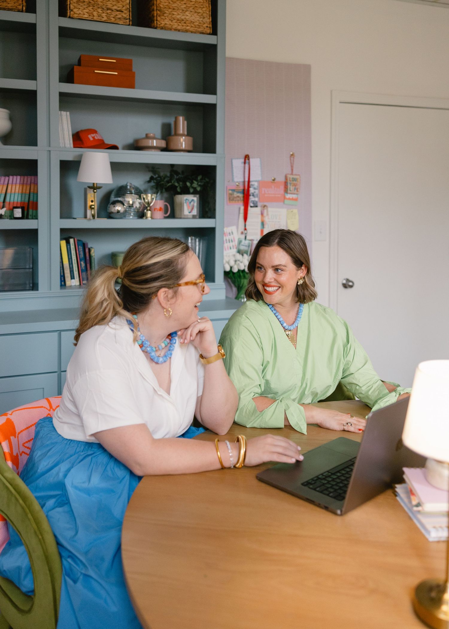 Two women smiling while working together on a laptop at a desk in a colorfully decorated office space.