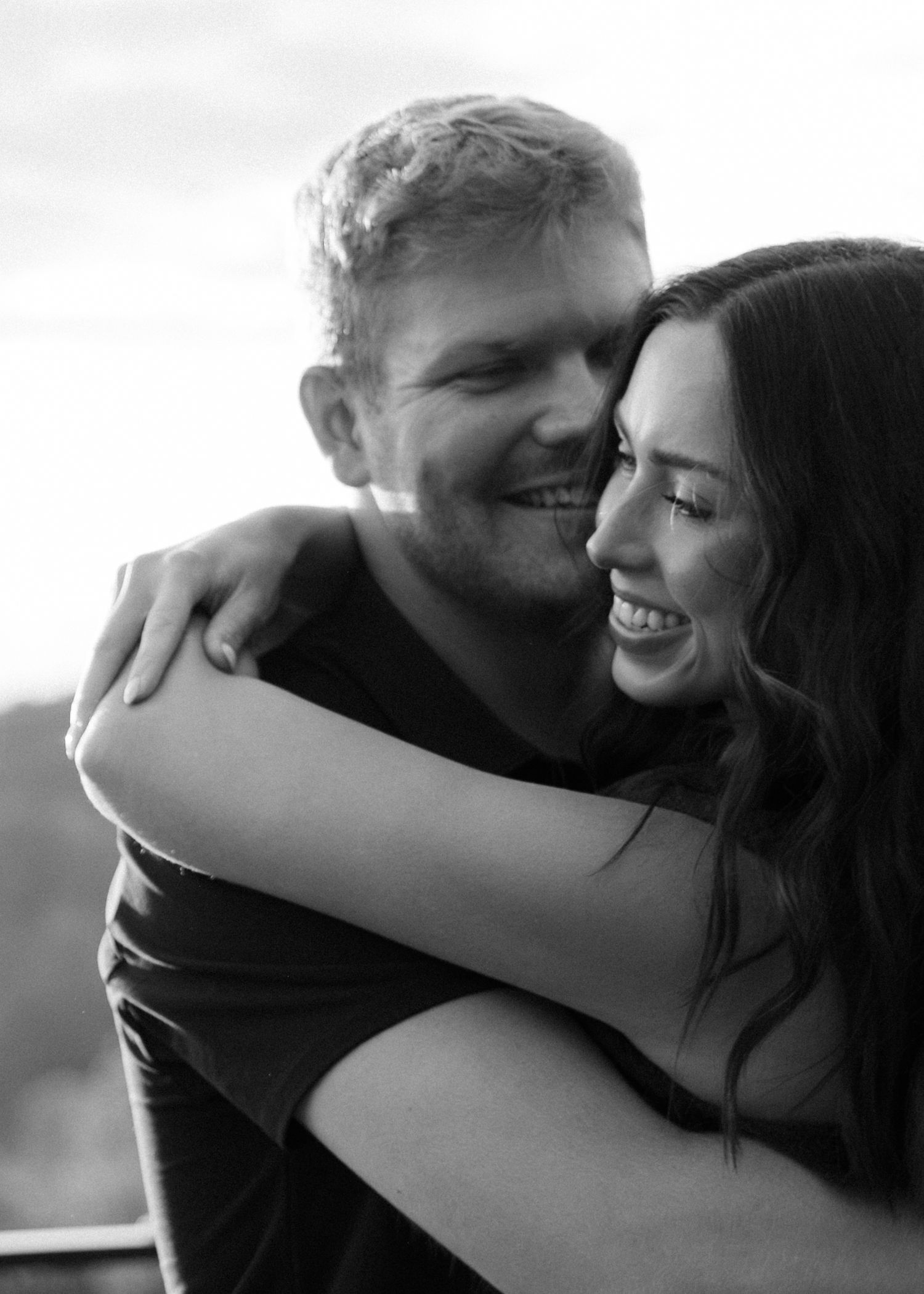 Black and white photo of a smiling couple embracing closely outdoors, the woman with long dark hair laughing as the man nuzzles her cheek.