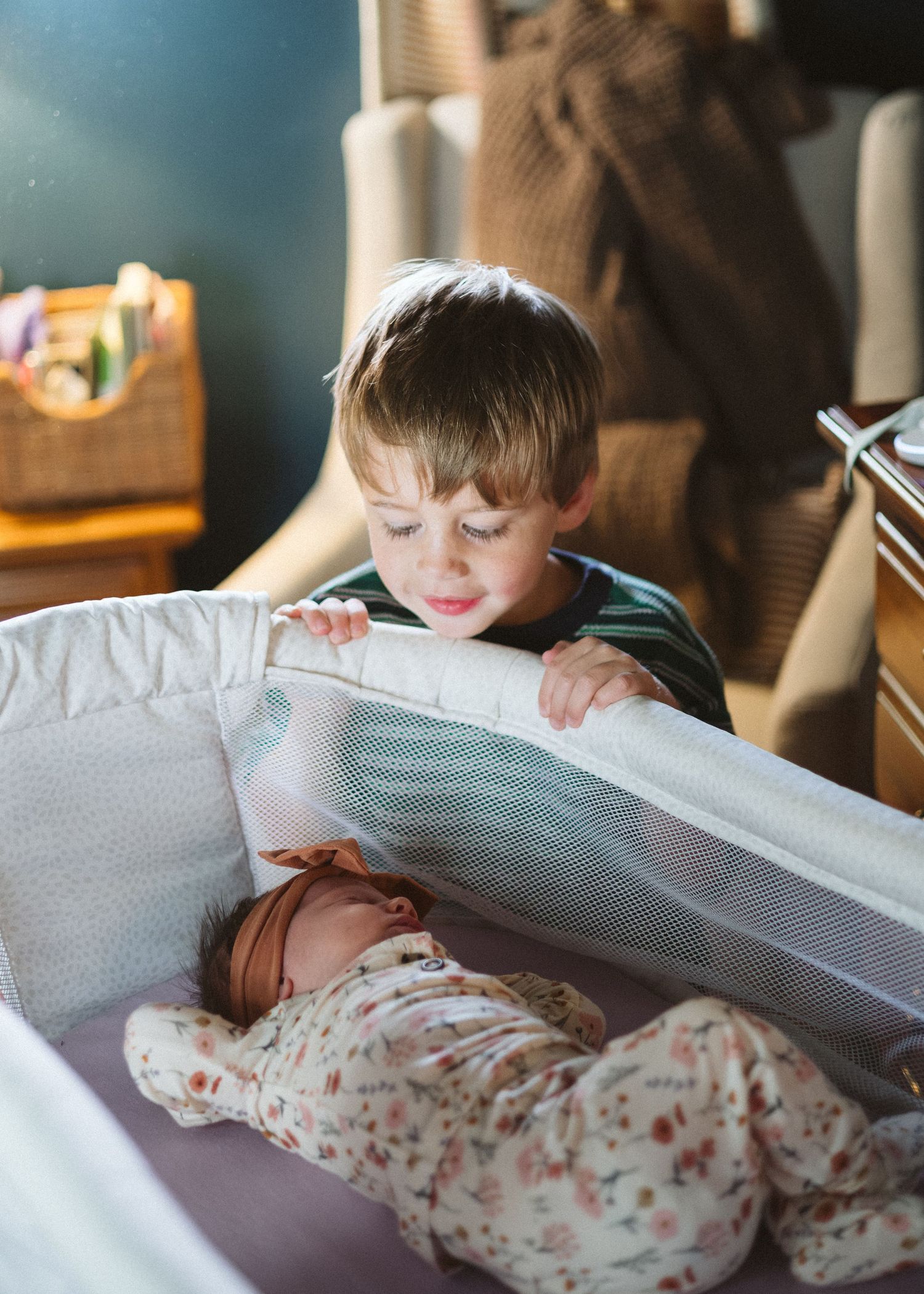 Curious young boy peers over the edge of a bassinet at a sleeping newborn baby in a floral outfit and headband.