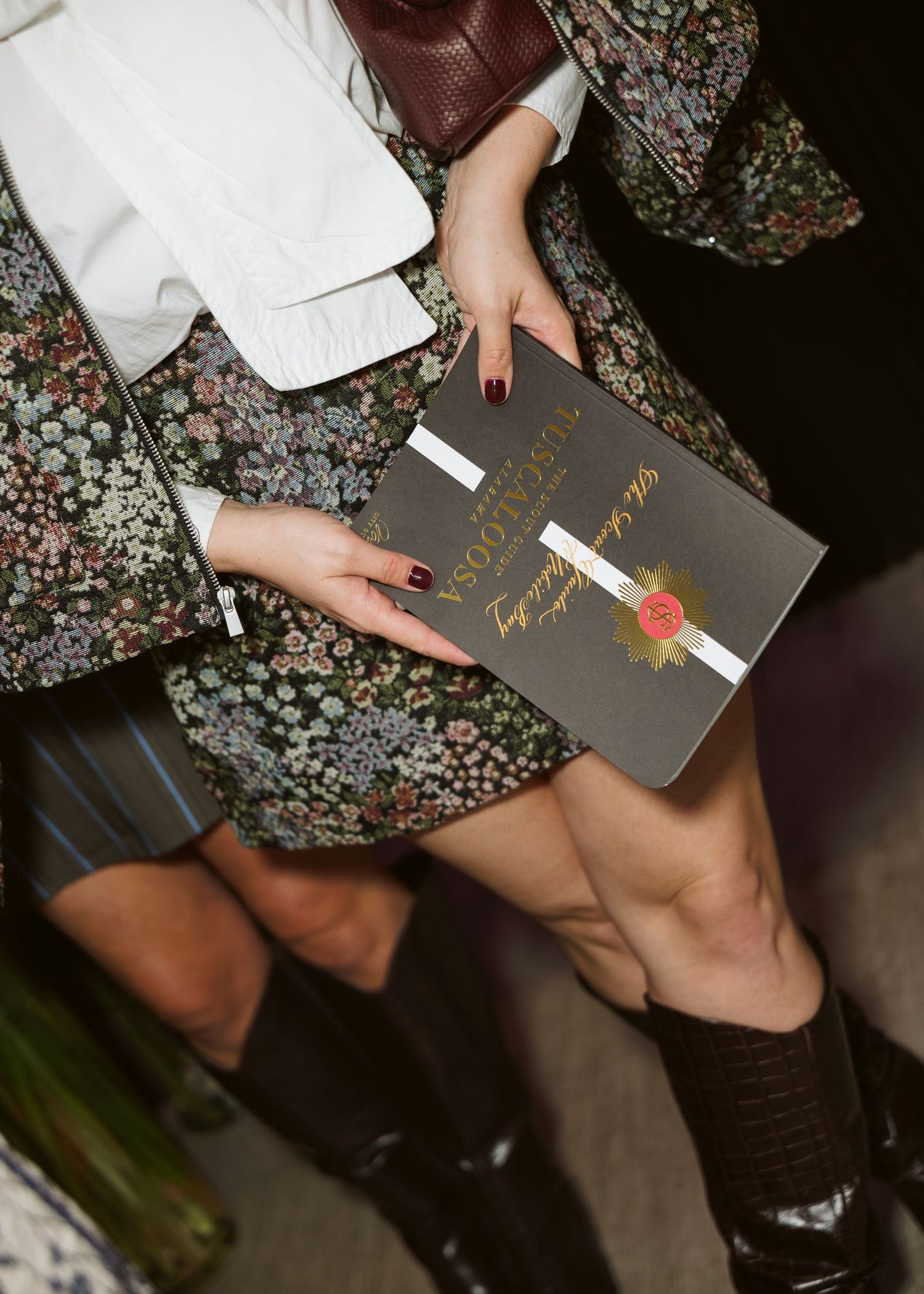 Woman in a floral mini dress, white jacket and black croc-embossed boots holds an elegant gray invitation sealed with a red wax stamp.
