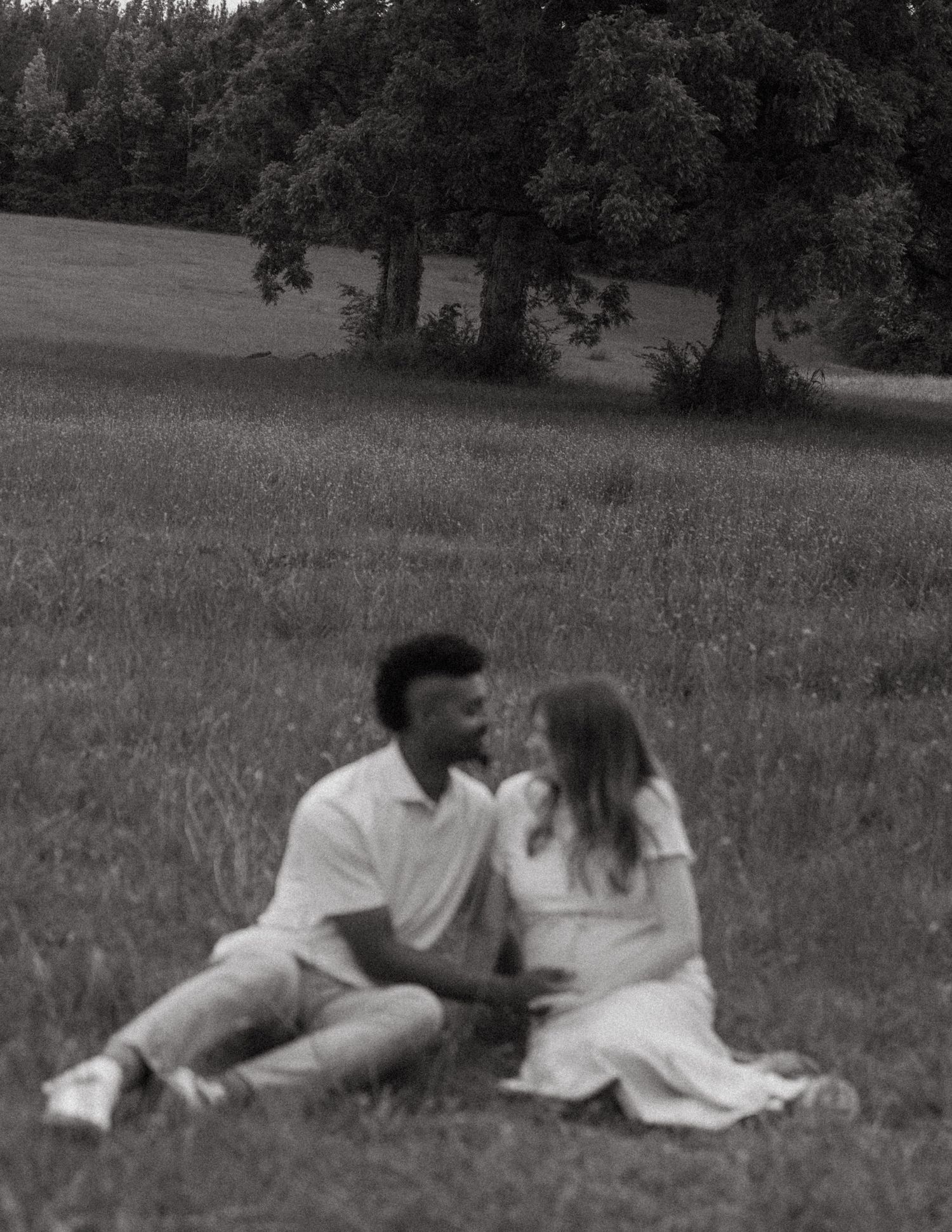 Black and white photo of expecting couple sitting together in a lush grassy field surrounded by trees.