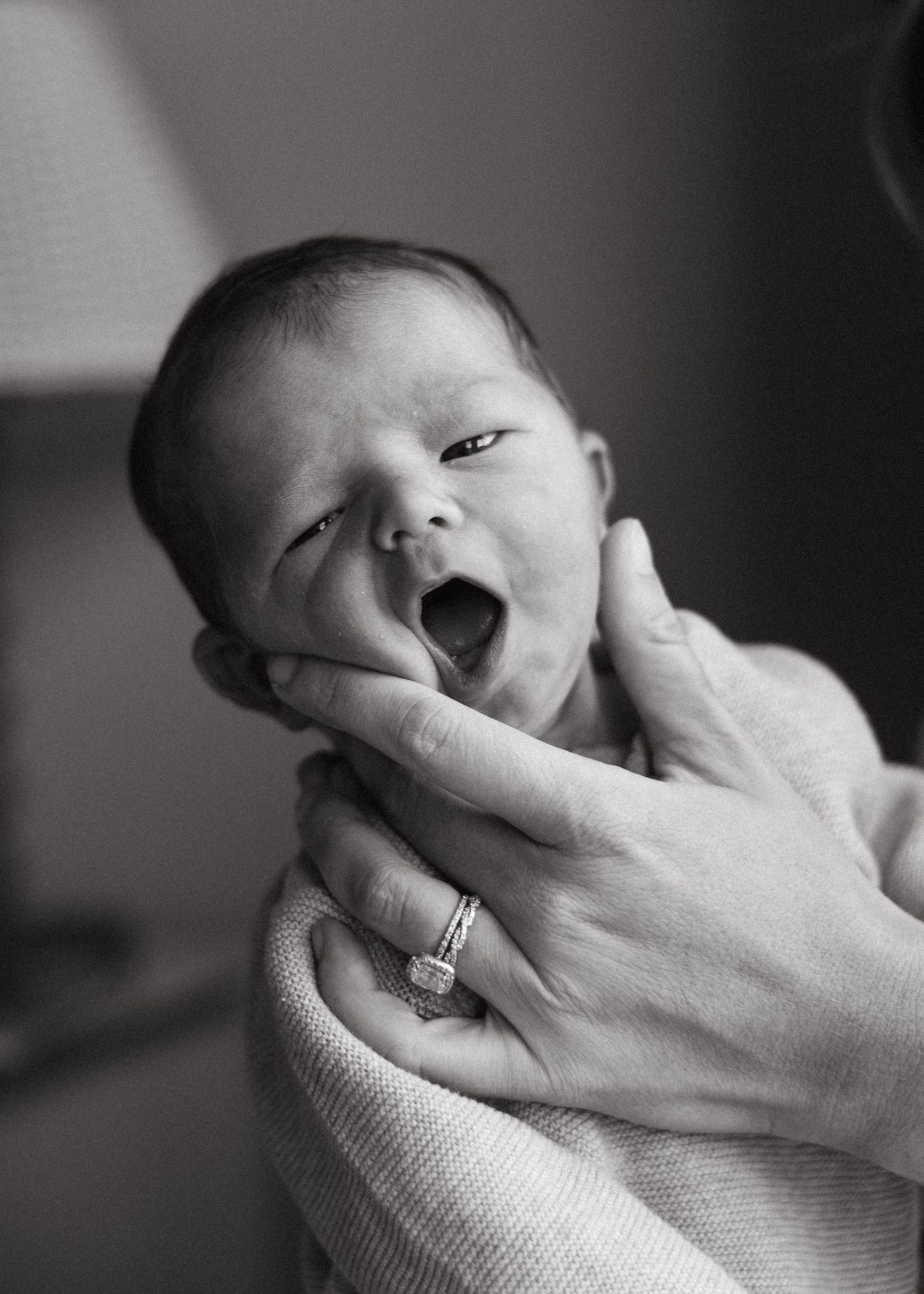 Black and white photo of yawning newborn baby cradled in mother's hands with diamond ring.