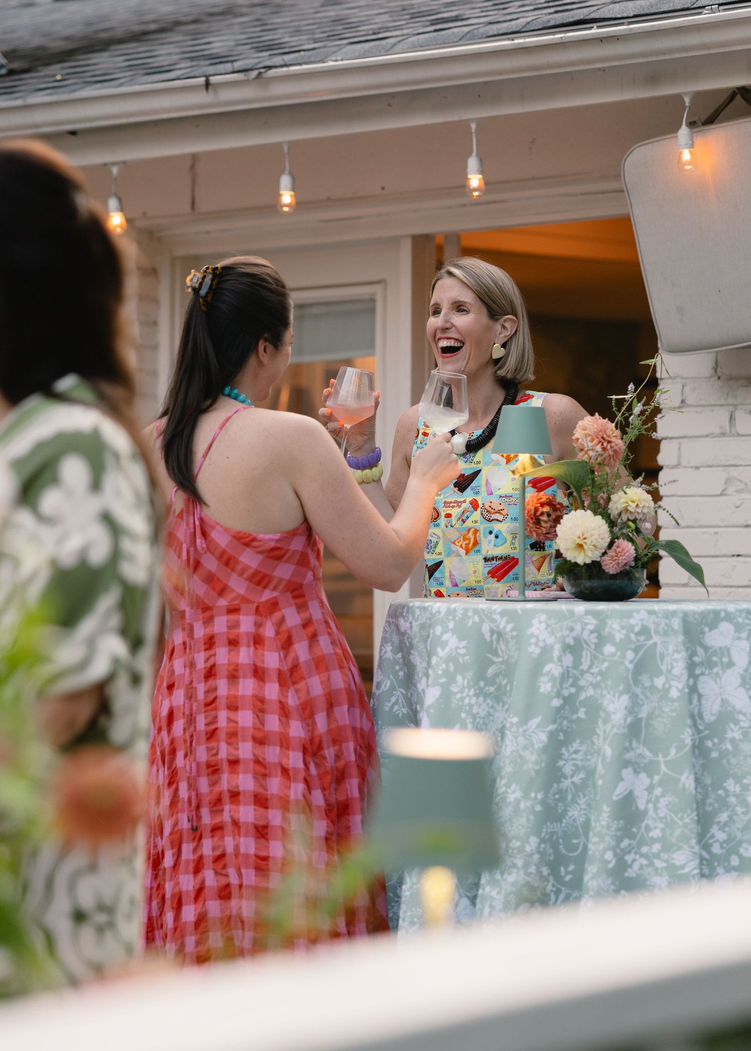 Two women laughing and toasting with wine glasses at an outdoor evening garden party with string lights.