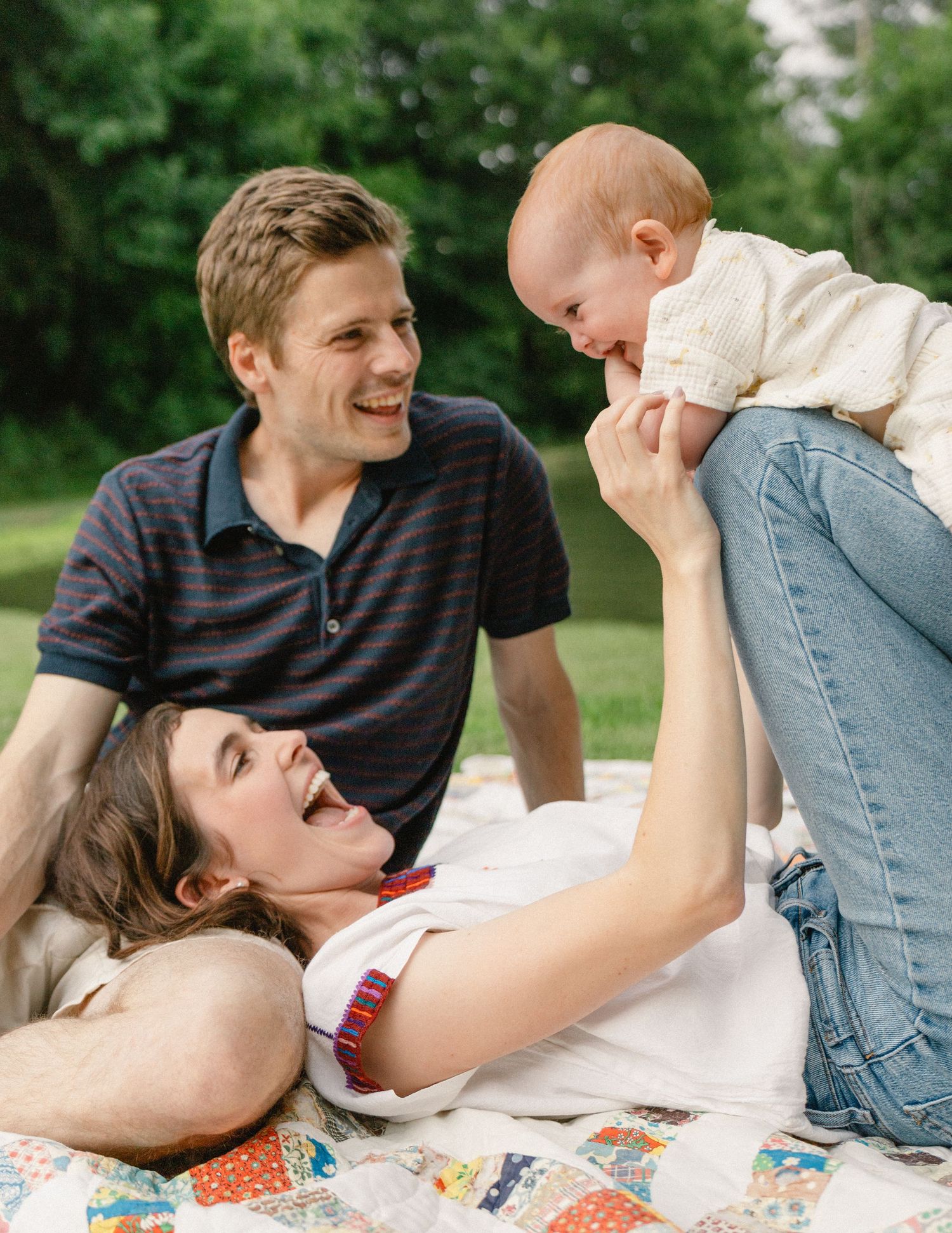 Happy family relaxing on a quilt outdoors with mother holding smiling baby in air.