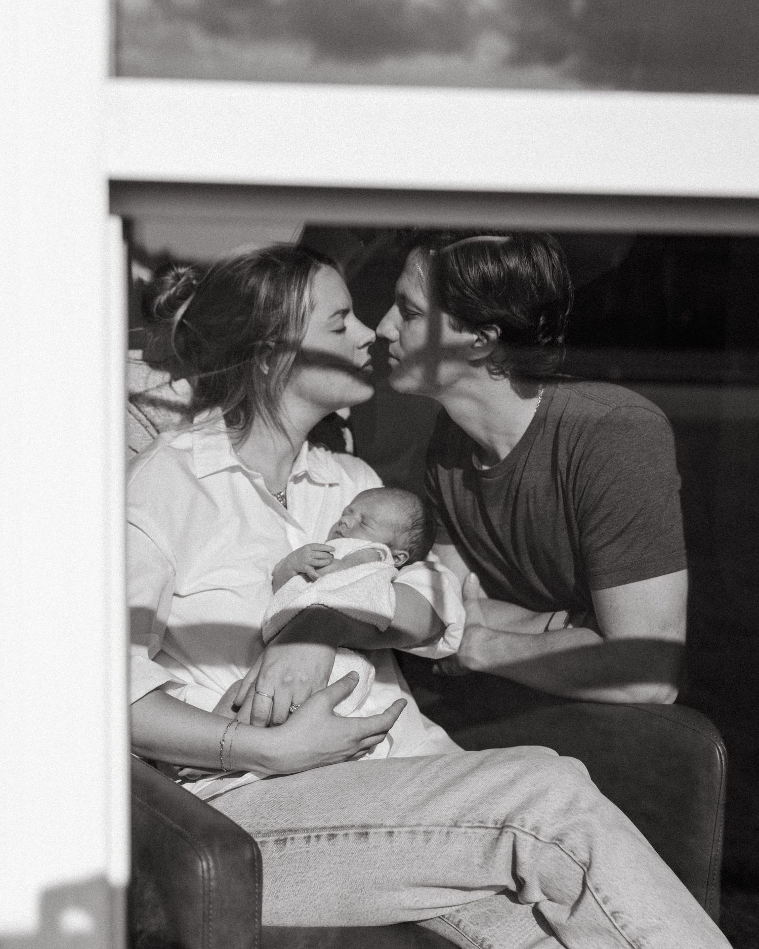 Black and white photo of couple kissing while cradling newborn baby, sitting together in warm natural light.