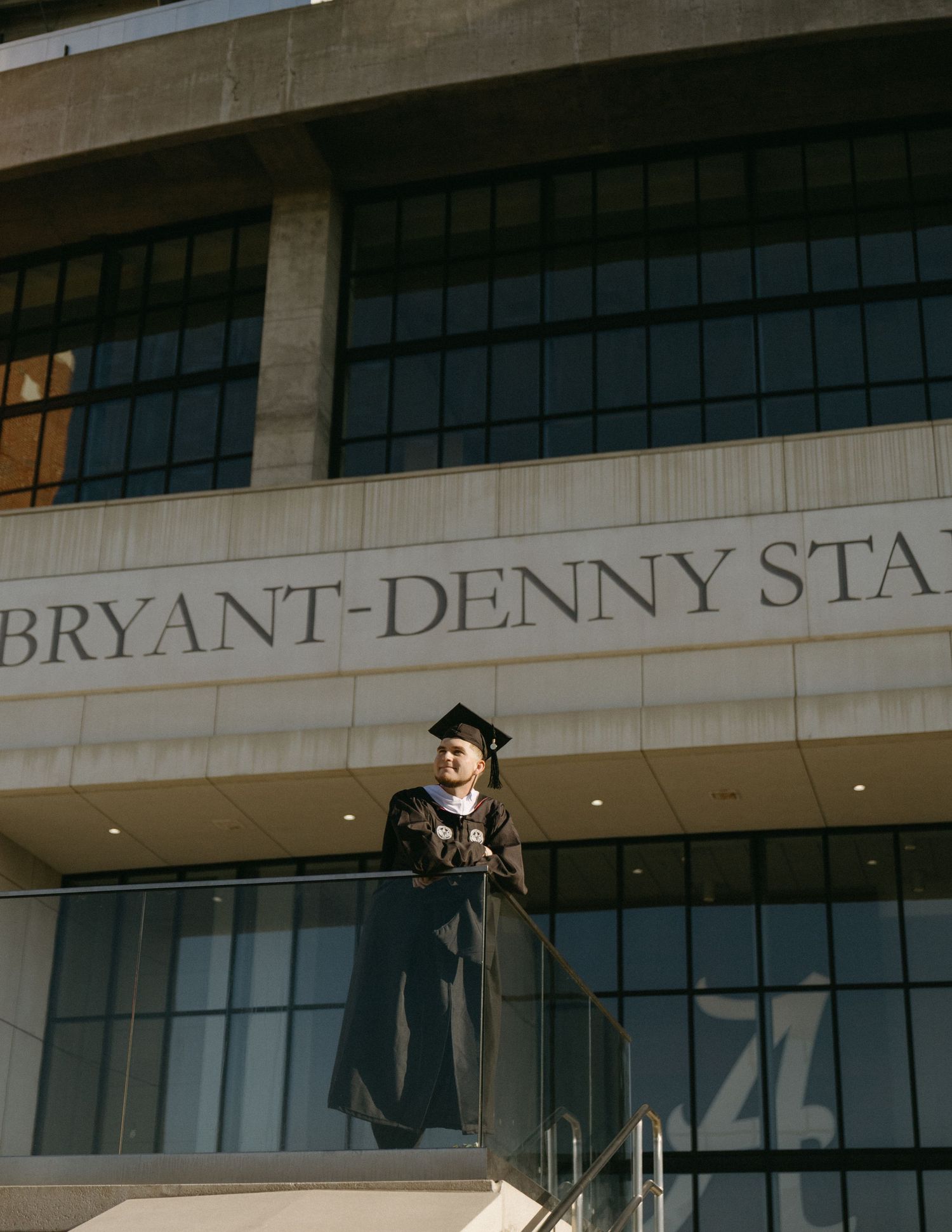 Graduate in cap and gown standing on balcony of Bryant-Denny Stadium in warm sunlight.