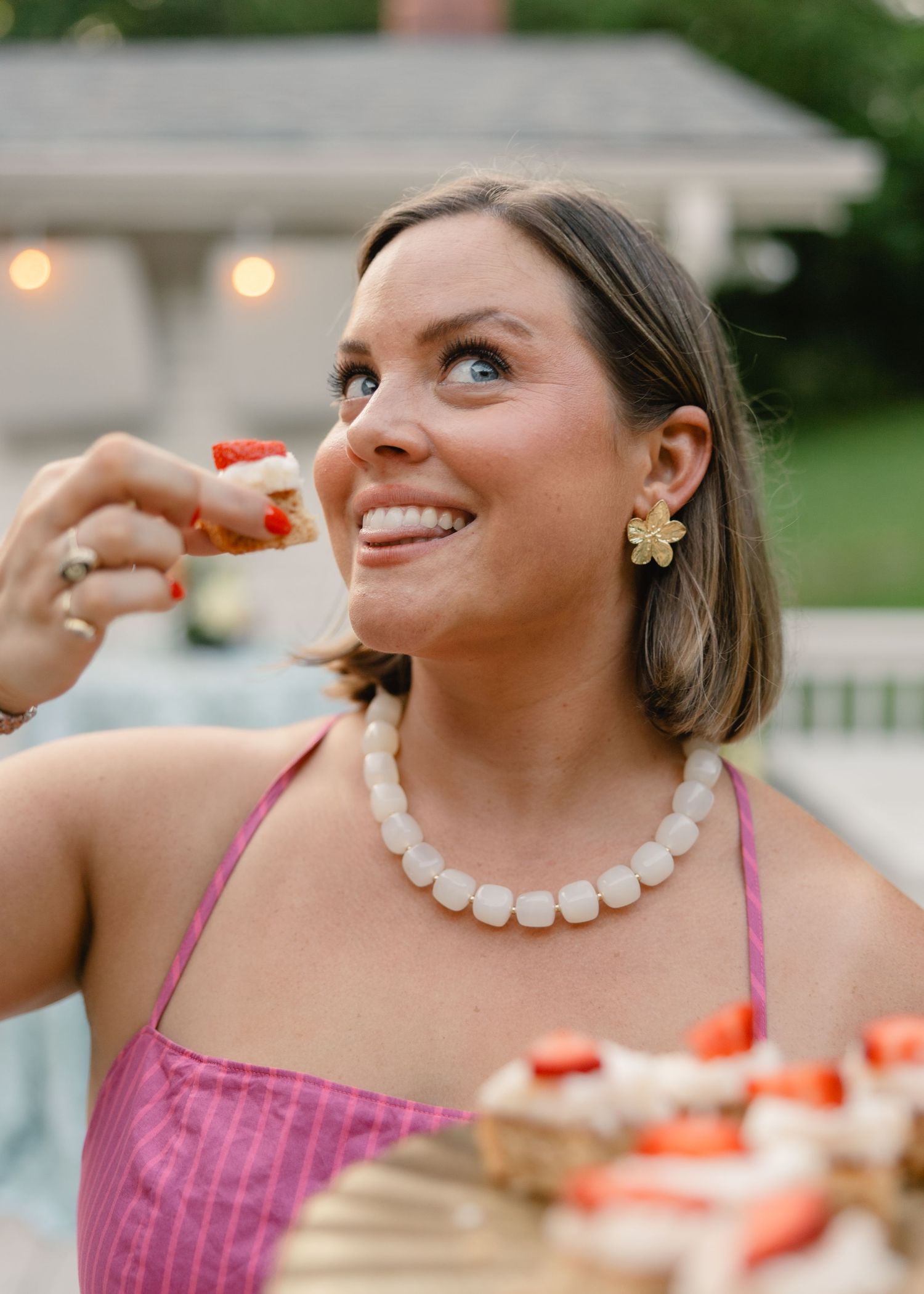 Woman in pink dress eating strawberry shortcake at outdoor party with string lights.