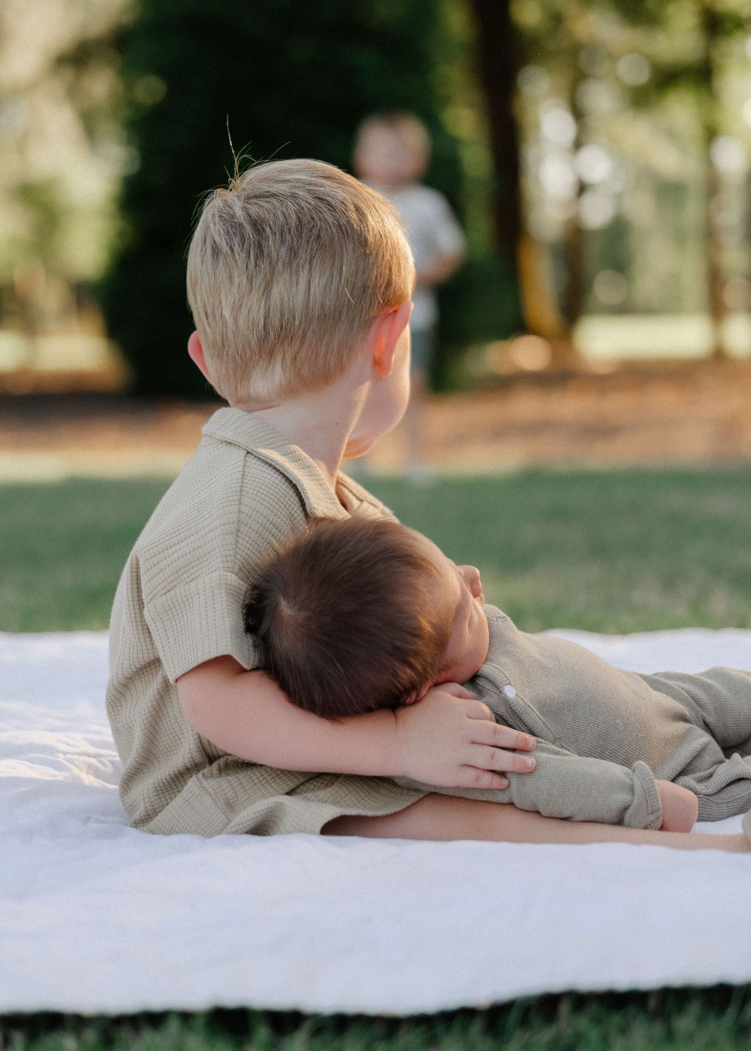 Young boy gently cradling a sleeping newborn sibling on a white blanket outdoors.