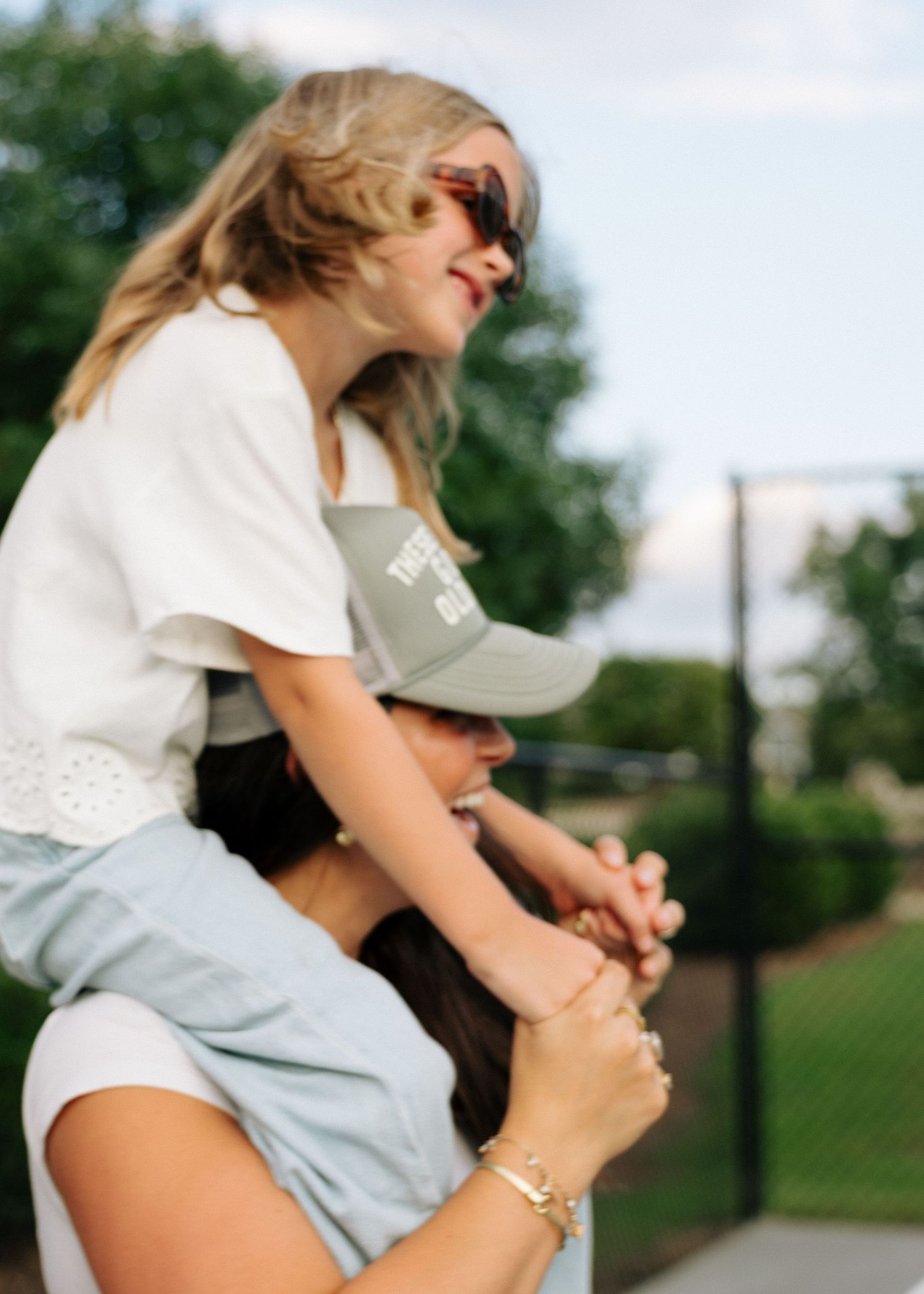 A girl in sunglasses sits on a womans shoulders while holding her hands outdoors.