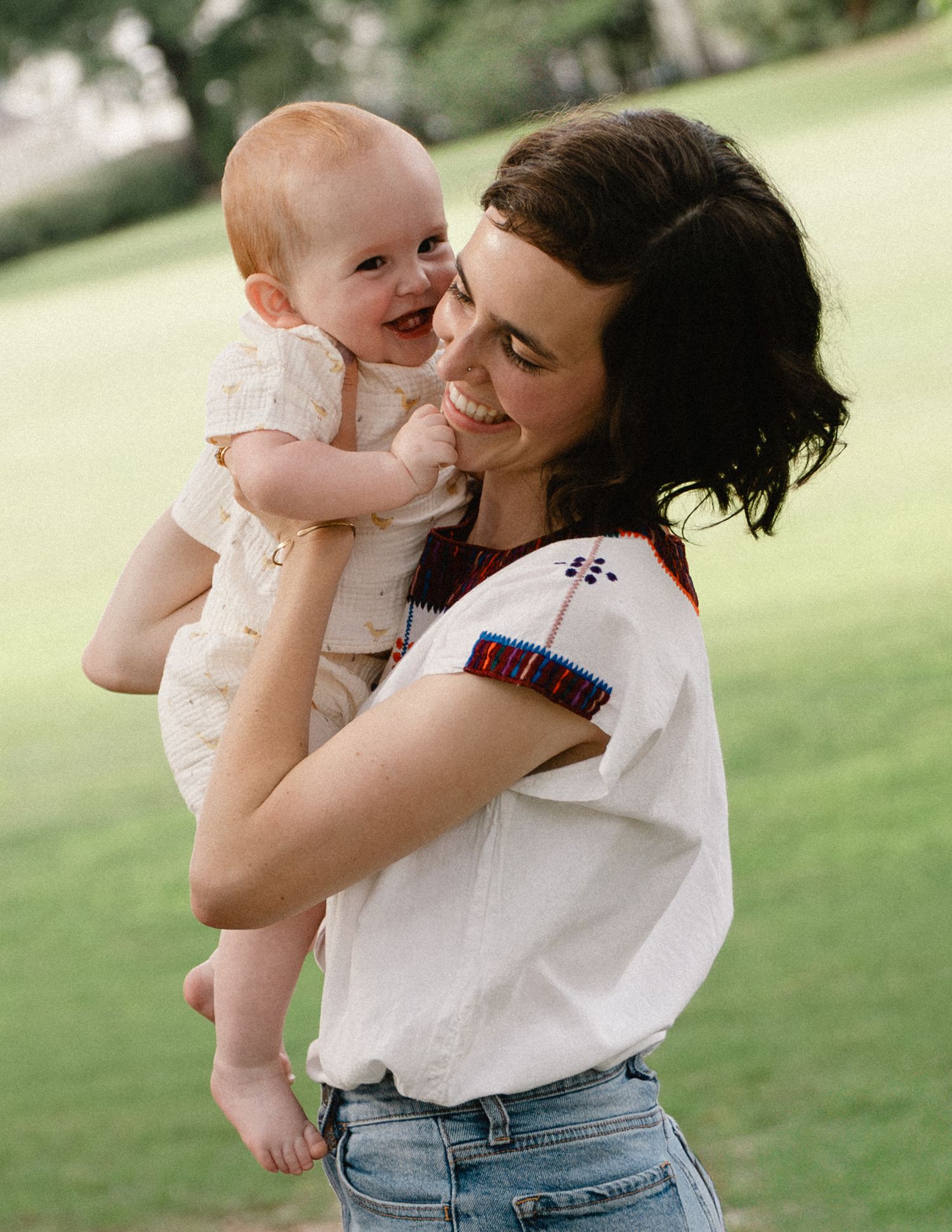 A smiling mother holds her happy baby close in a green park setting.