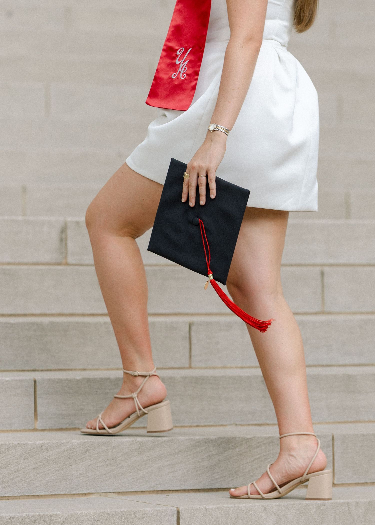Graduate holds black cap with red tassel wearing white dress and nude heels on stone steps.