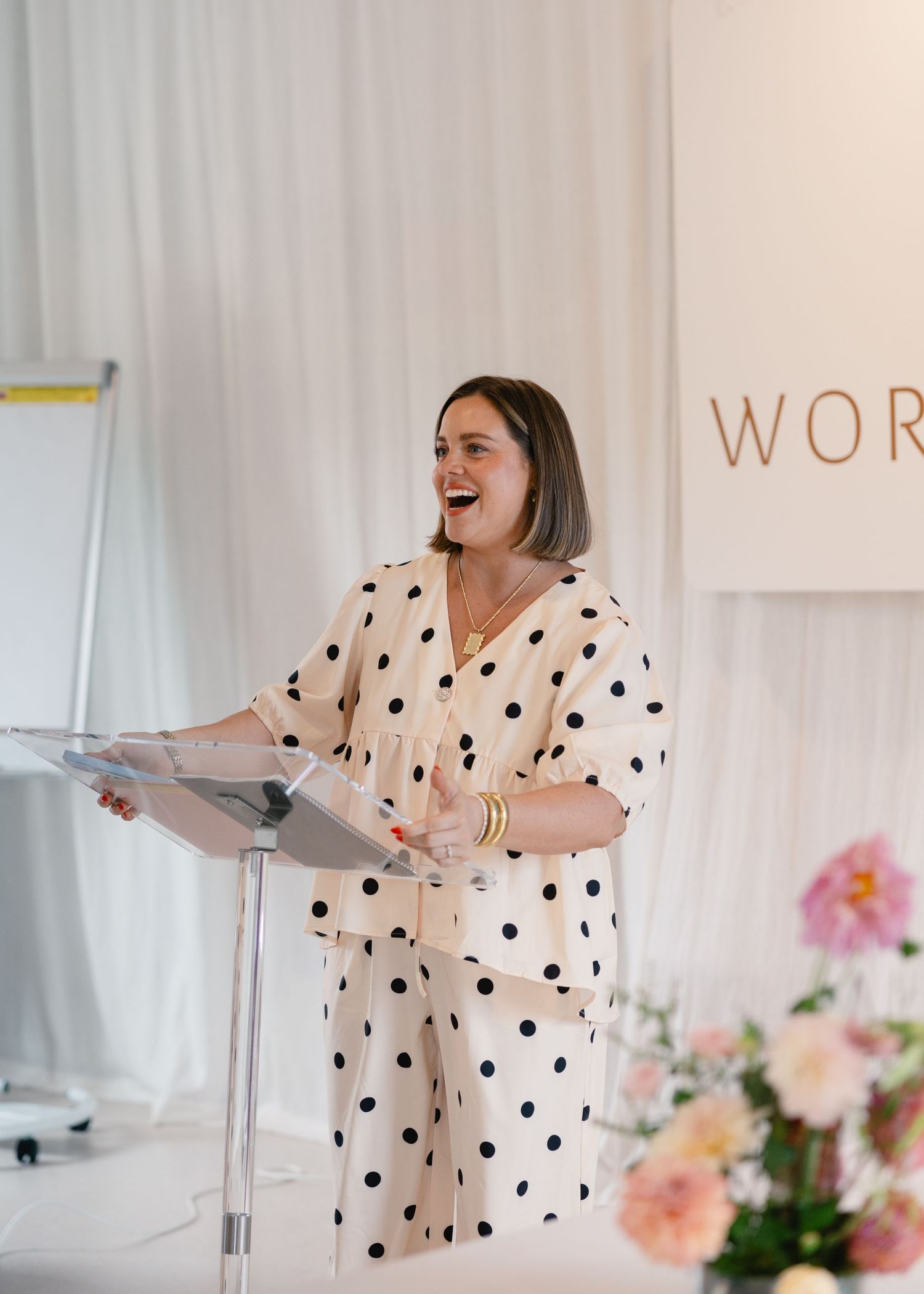 Woman in polka dot outfit speaking at podium during Workshop event with floral decor.