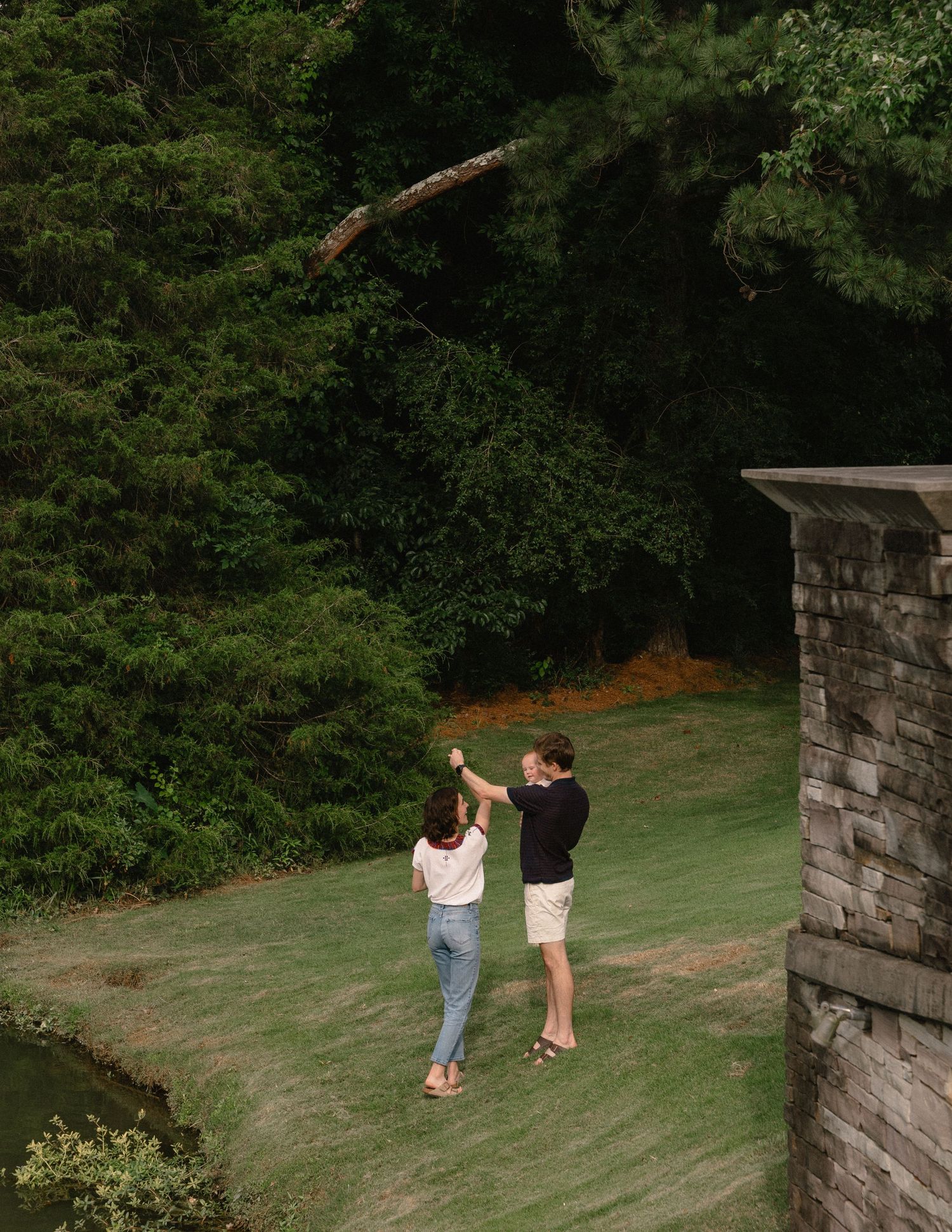 Couple dancing together on a green lawn surrounded by lush trees near a stone wall.