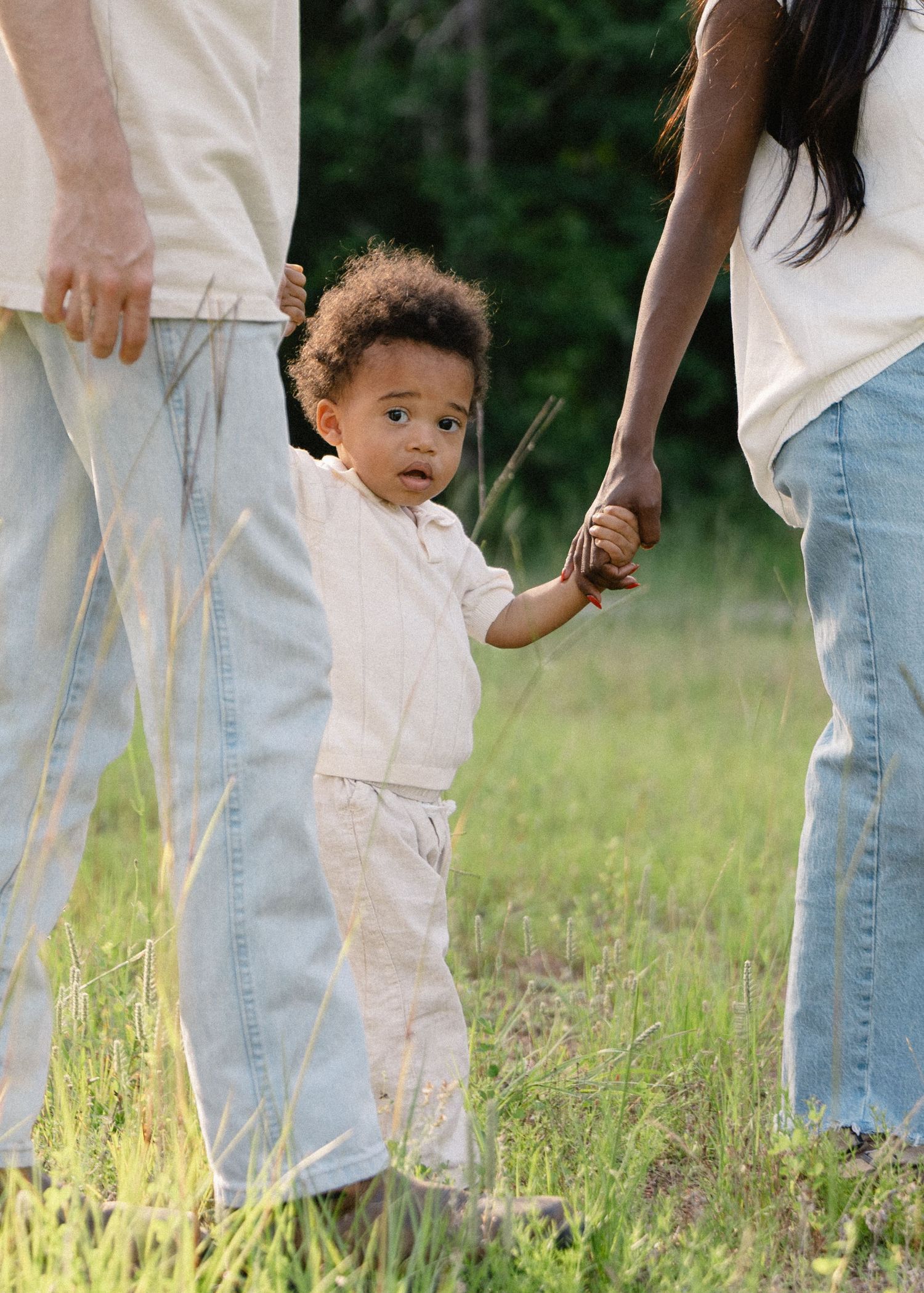 A toddler holds hands with parents while walking through a sunny green meadow.