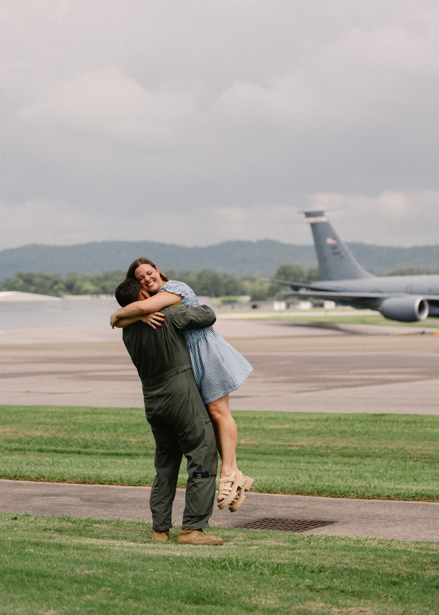 A couple shares a joyful spinning embrace on a military airfield with a large aircraft visible in the background.