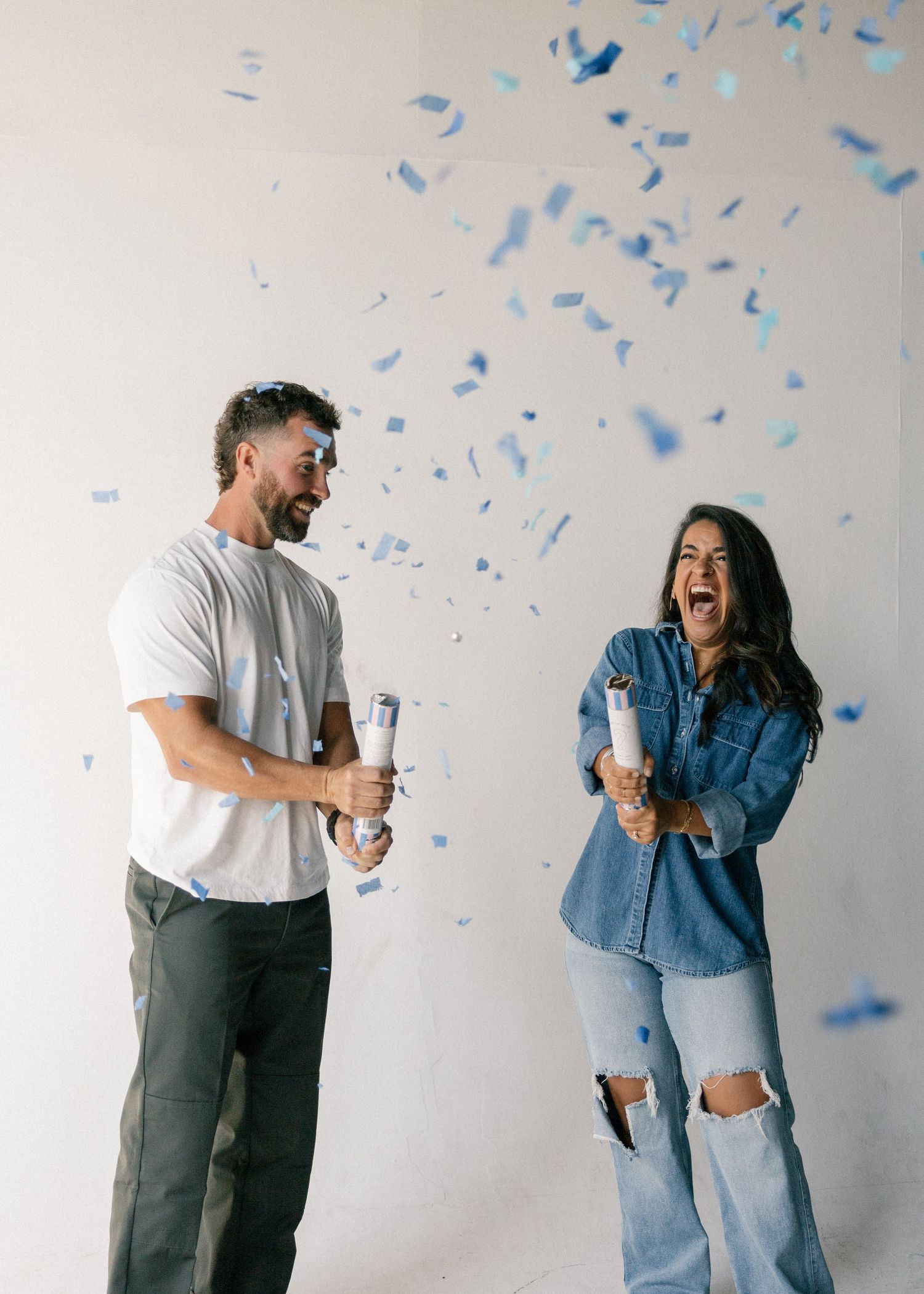 Couple laughing as blue confetti explodes from poppers during a baby boy gender reveal photoshoot.