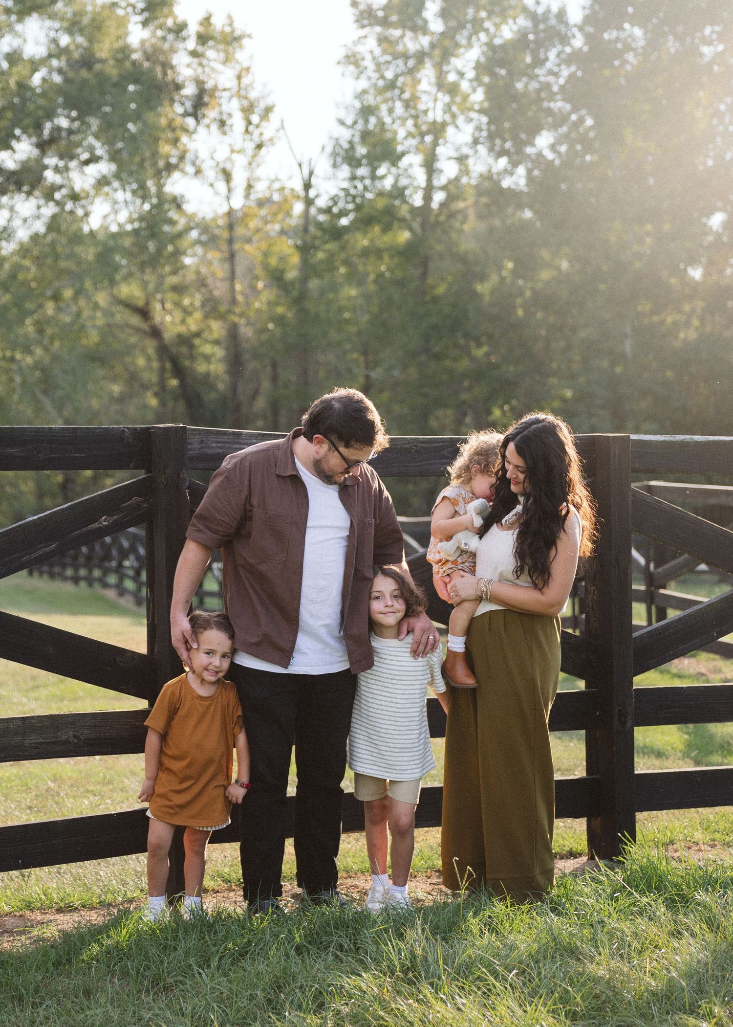Family of five poses together by a wooden fence in a sunlit grassy field, with parents and three young children smiling at golden hour.