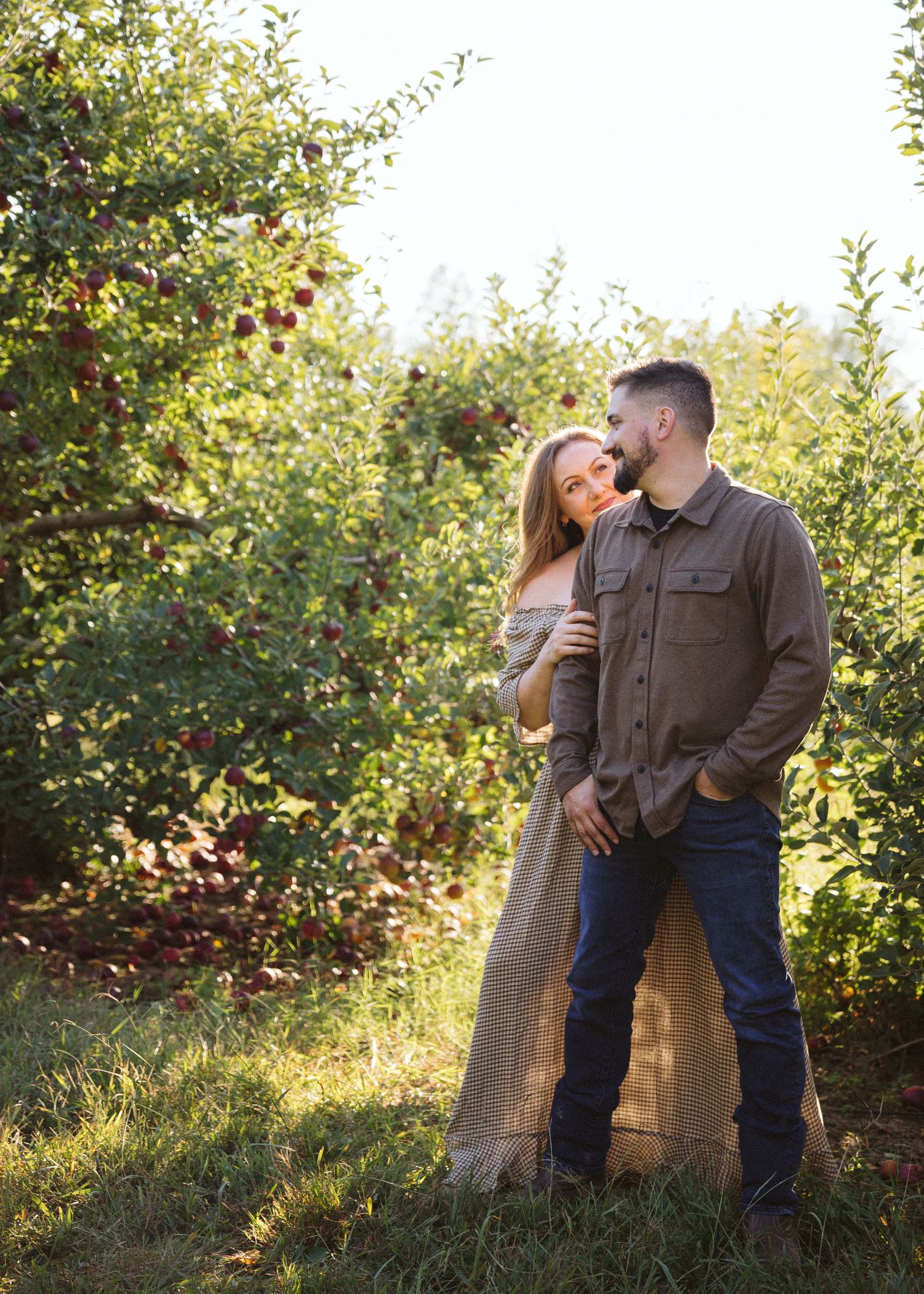 Couple embracing in a sunlit apple orchard, the woman in a flowing off-shoulder dress and the man in a button-up shirt and jeans.