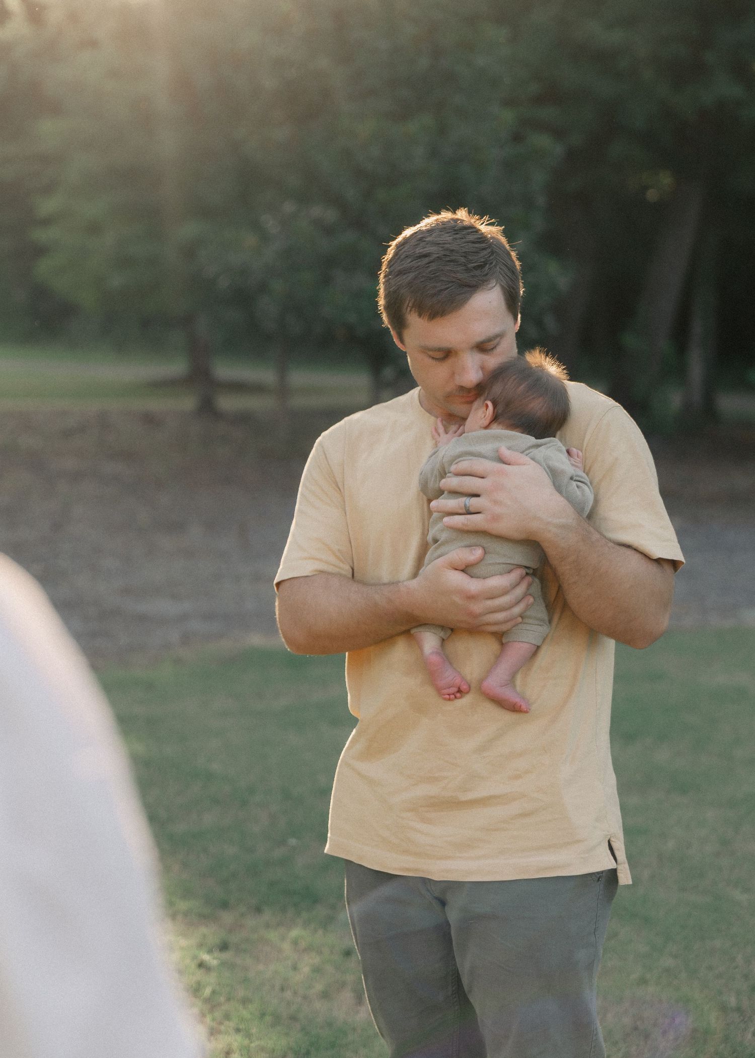 Father in yellow shirt lovingly cradling a newborn baby in a sunlit park.