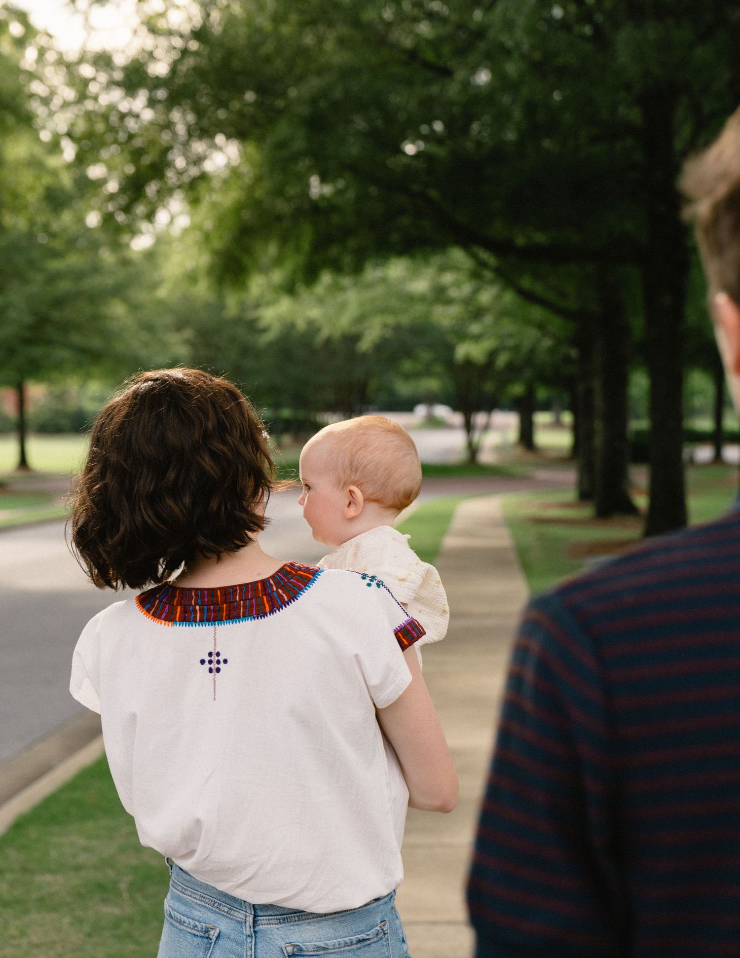 Mother carrying baby on her hip while walking down a tree-lined suburban street.