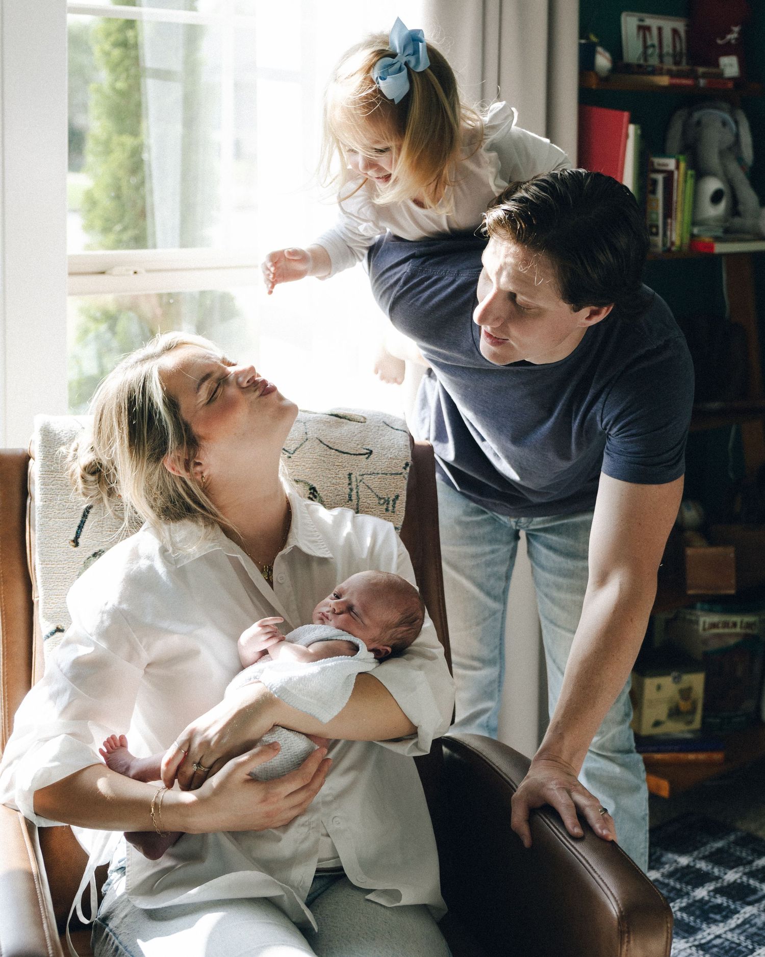 Family portrait with mother holding newborn, toddler daughter climbing on father near bright window.