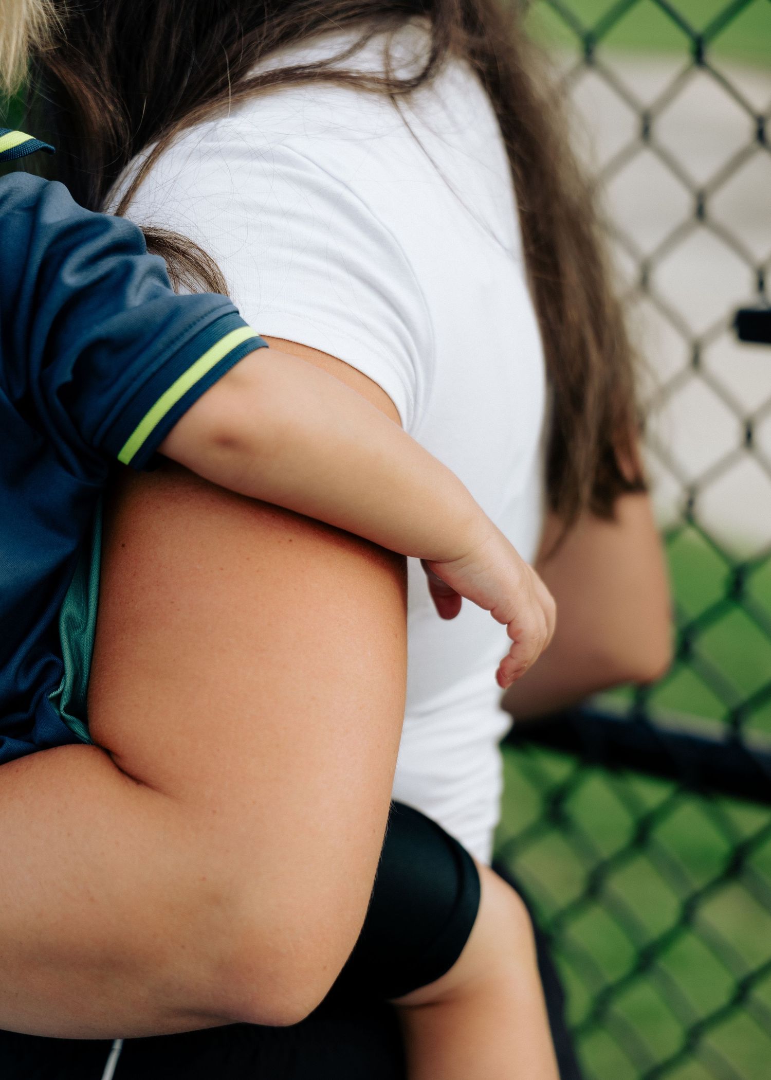 A woman in a white shirt holds a toddler in a blue jersey near a chain-link fence.