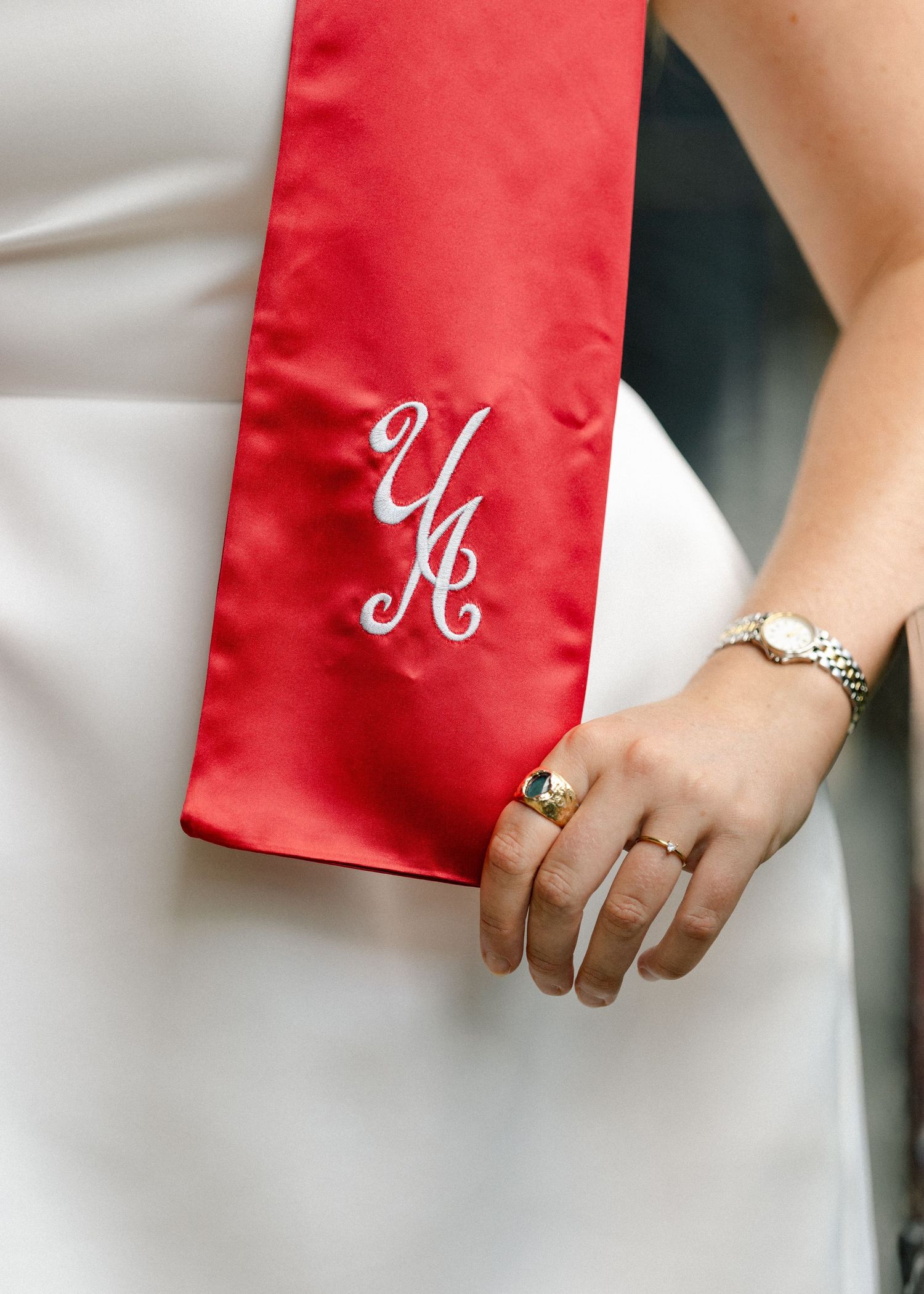 Close-up of red satin graduation stole with monogram YA, worn over white dress with ring and watch.