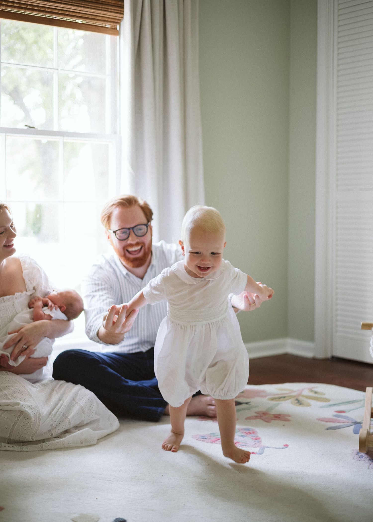 Smiling toddler in a white outfit takes wobbly first steps on a floral rug while a bearded dad and mom holding a newborn watch nearby.