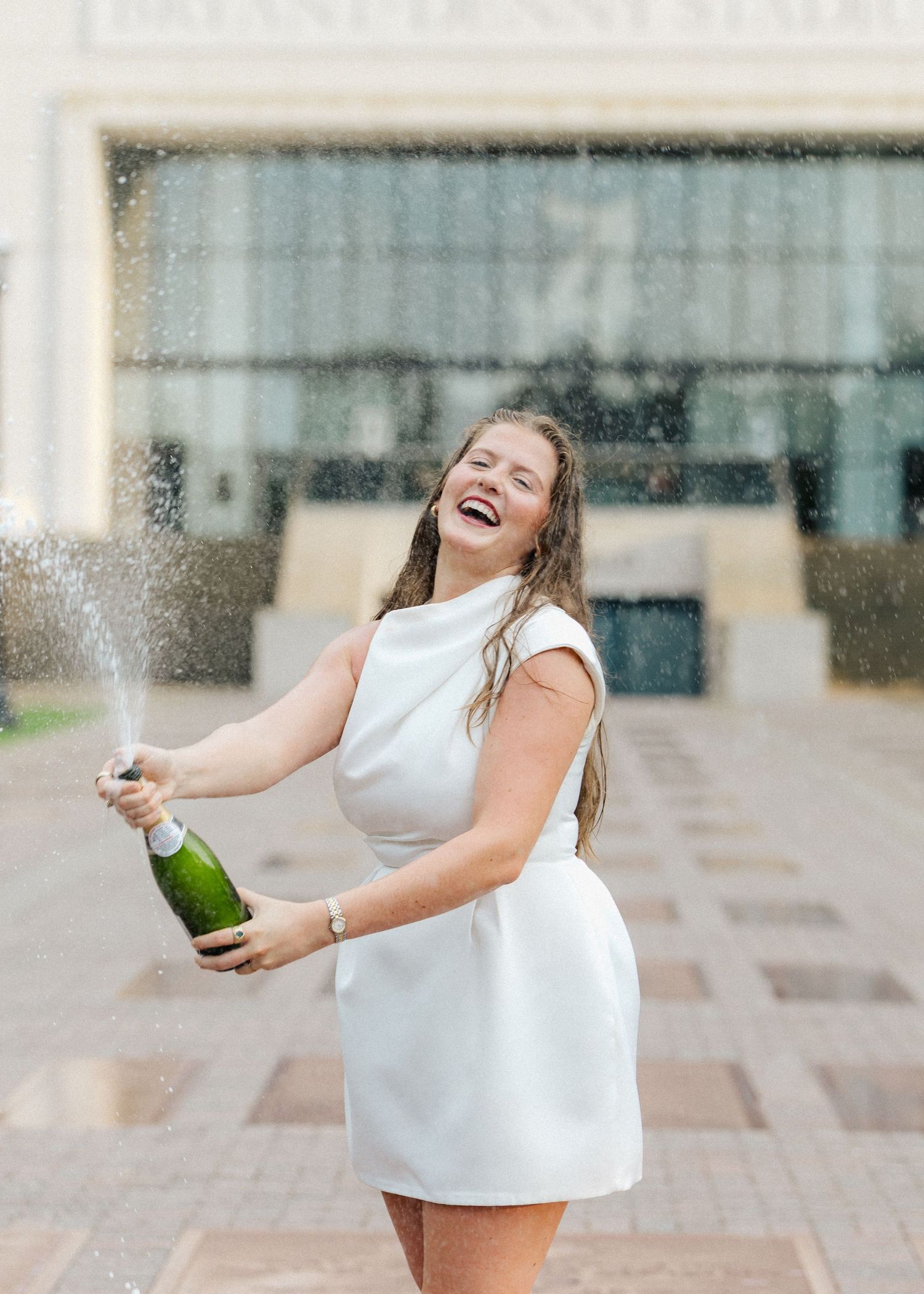 Laughing graduate in white dress pops champagne bottle outside university building.