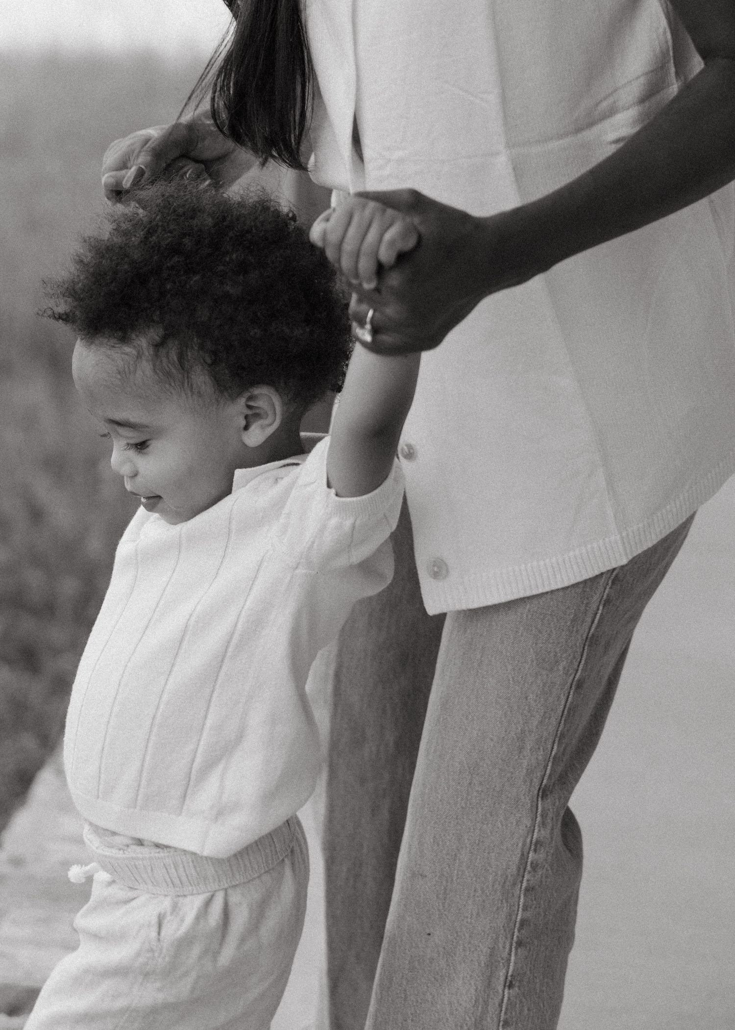 Black and white photo of a toddler holding hands with their mother while learning to walk outdoors.