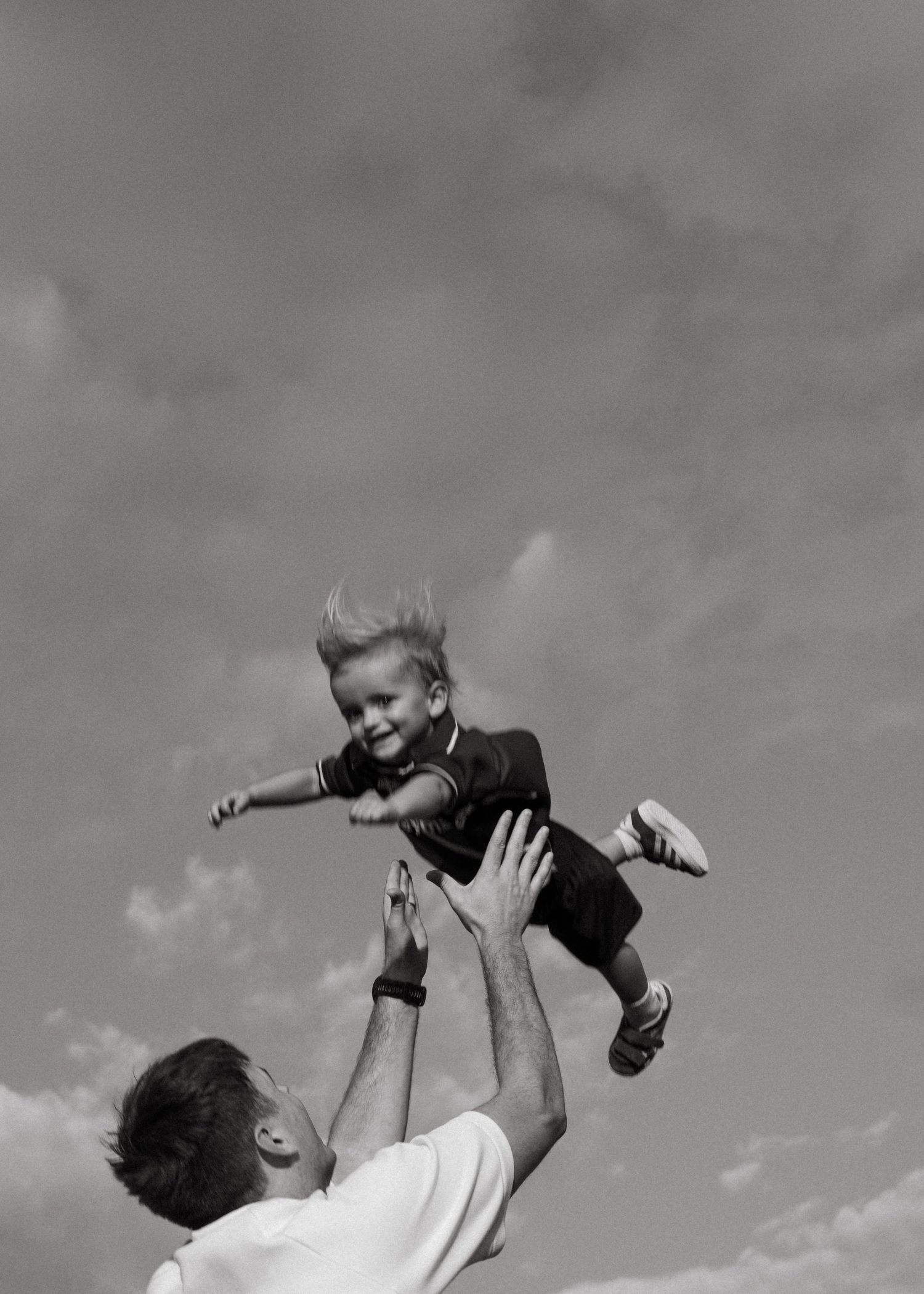 A black and white photo of a father tossing a laughing toddler high into the air against a cloudy sky.