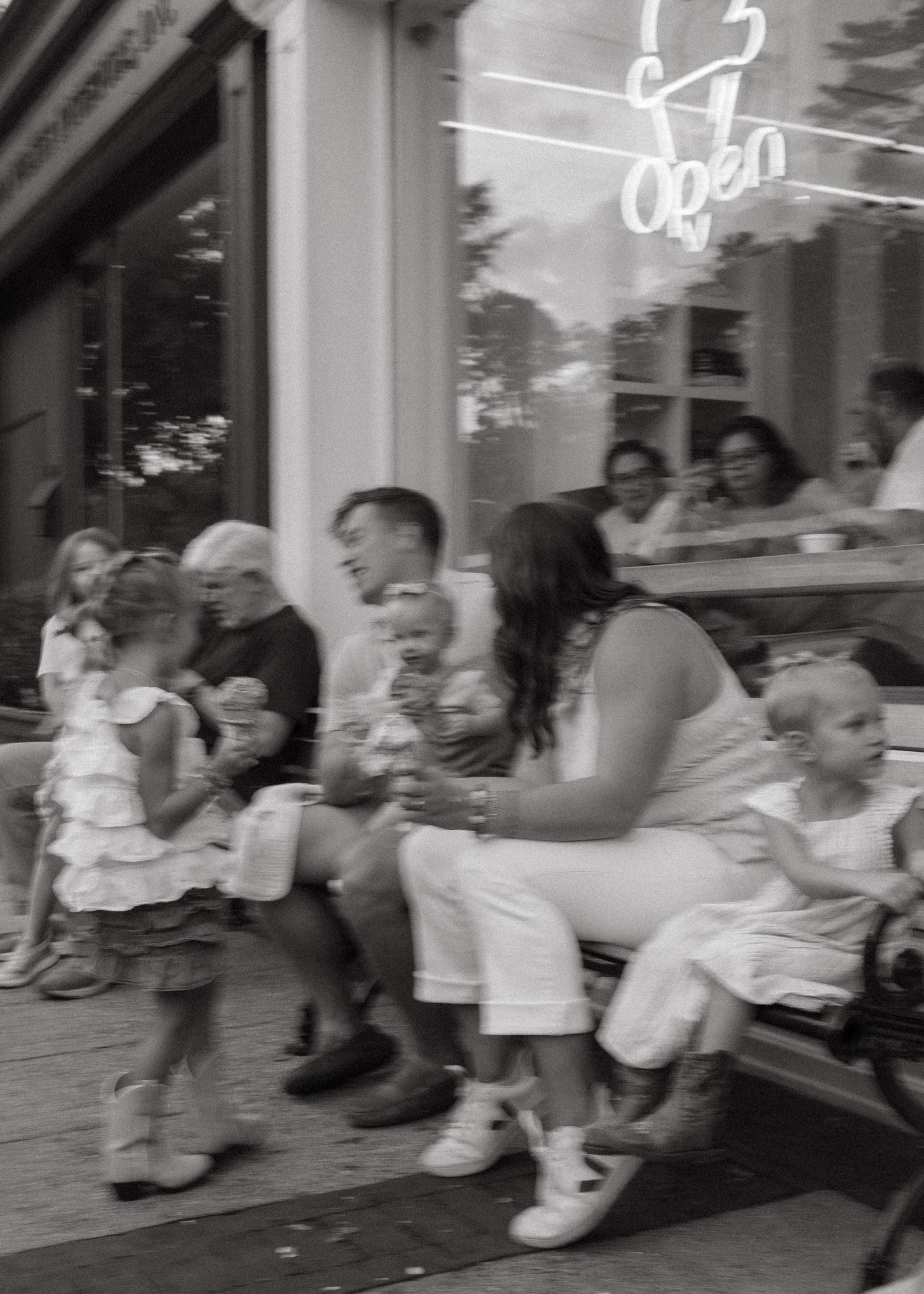 Black and white candid photo of a family with young children sitting outside an open storefront.